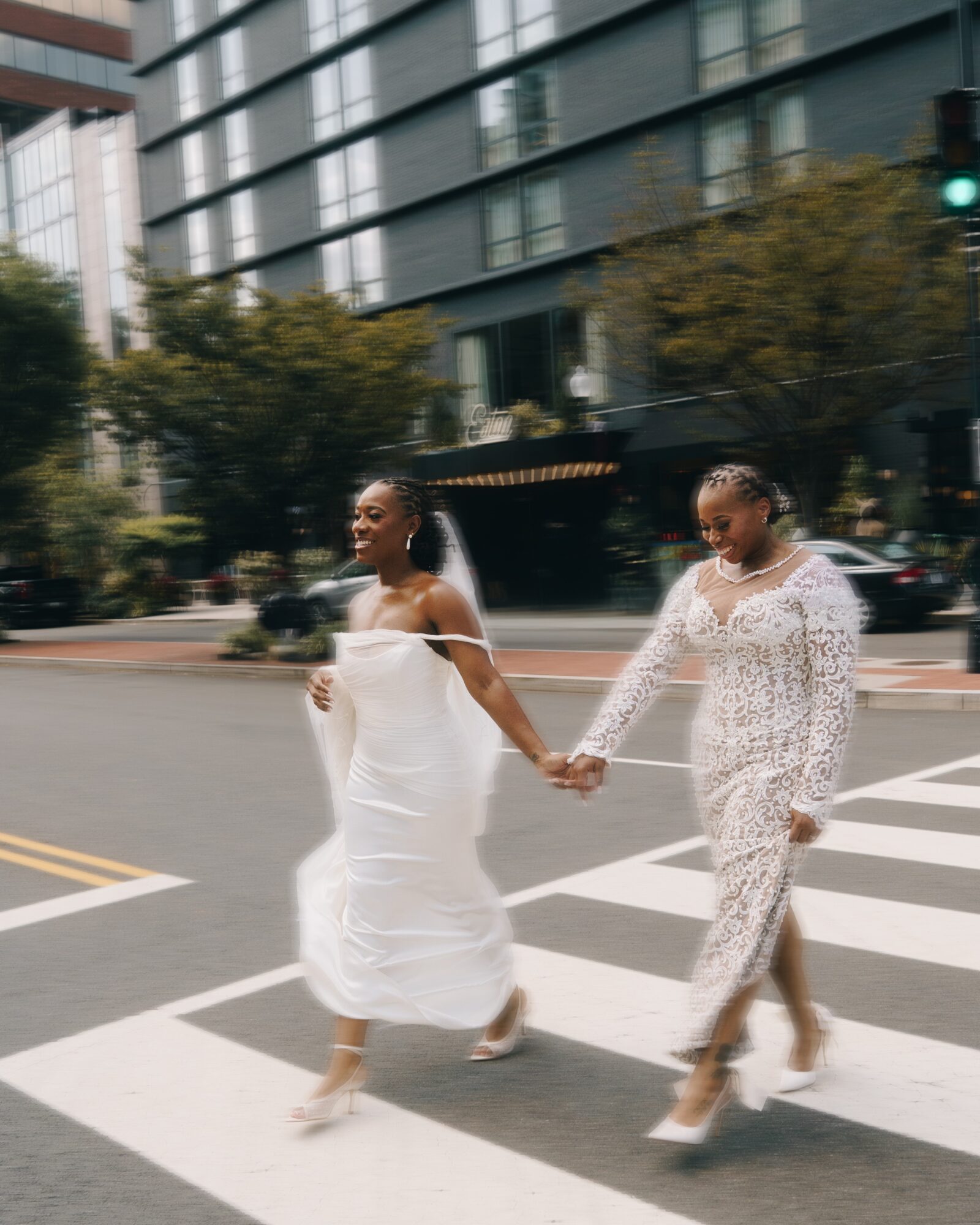 Two women holding hands and walking across a city crosswalk, smiling, with buildings and trees in the background.