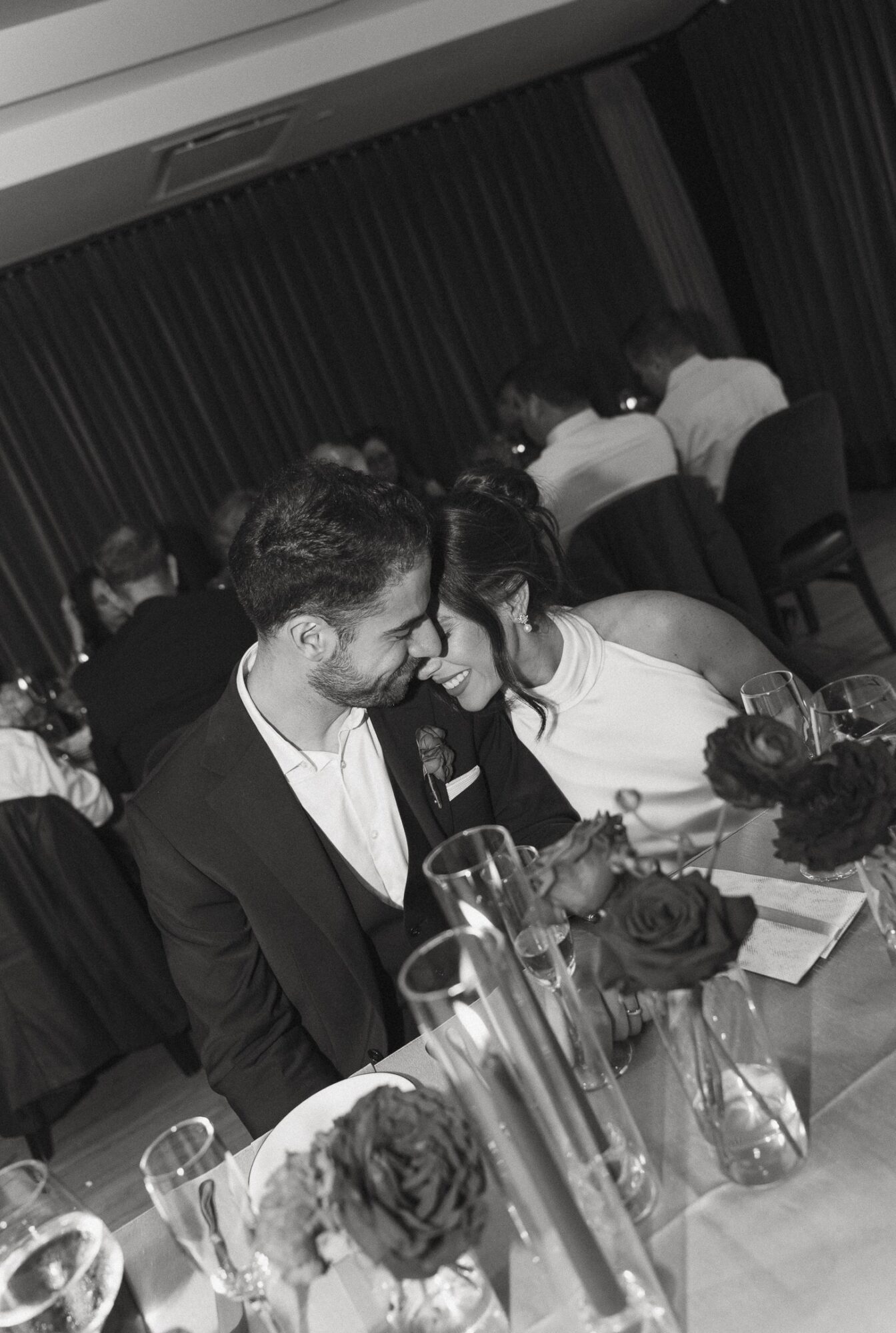 Two people sit close together at a table, smiling and looking at a phone, with flowers and glasses on the table.