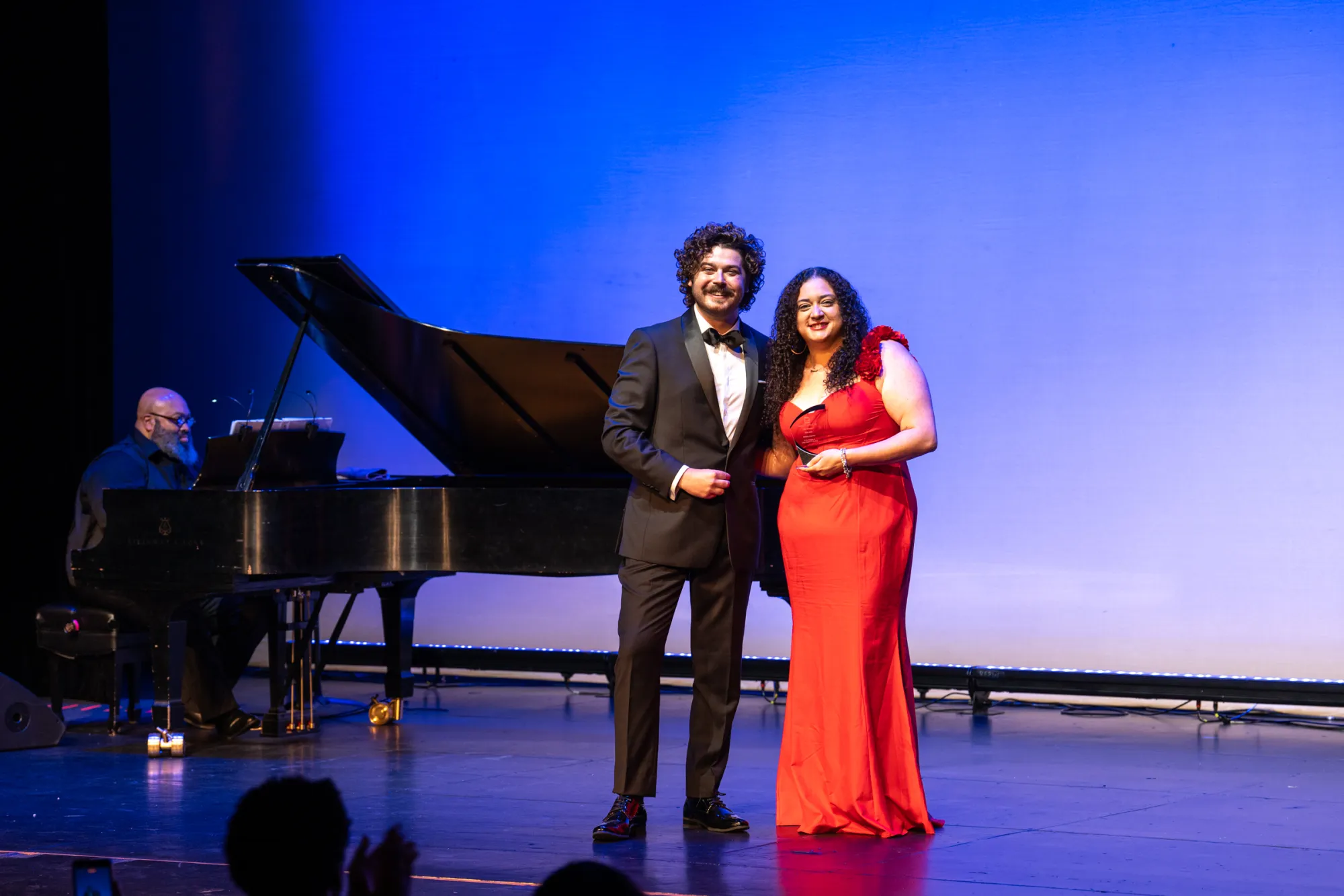 Two people in formal attire stand on stage with a pianist playing a grand piano in the background.