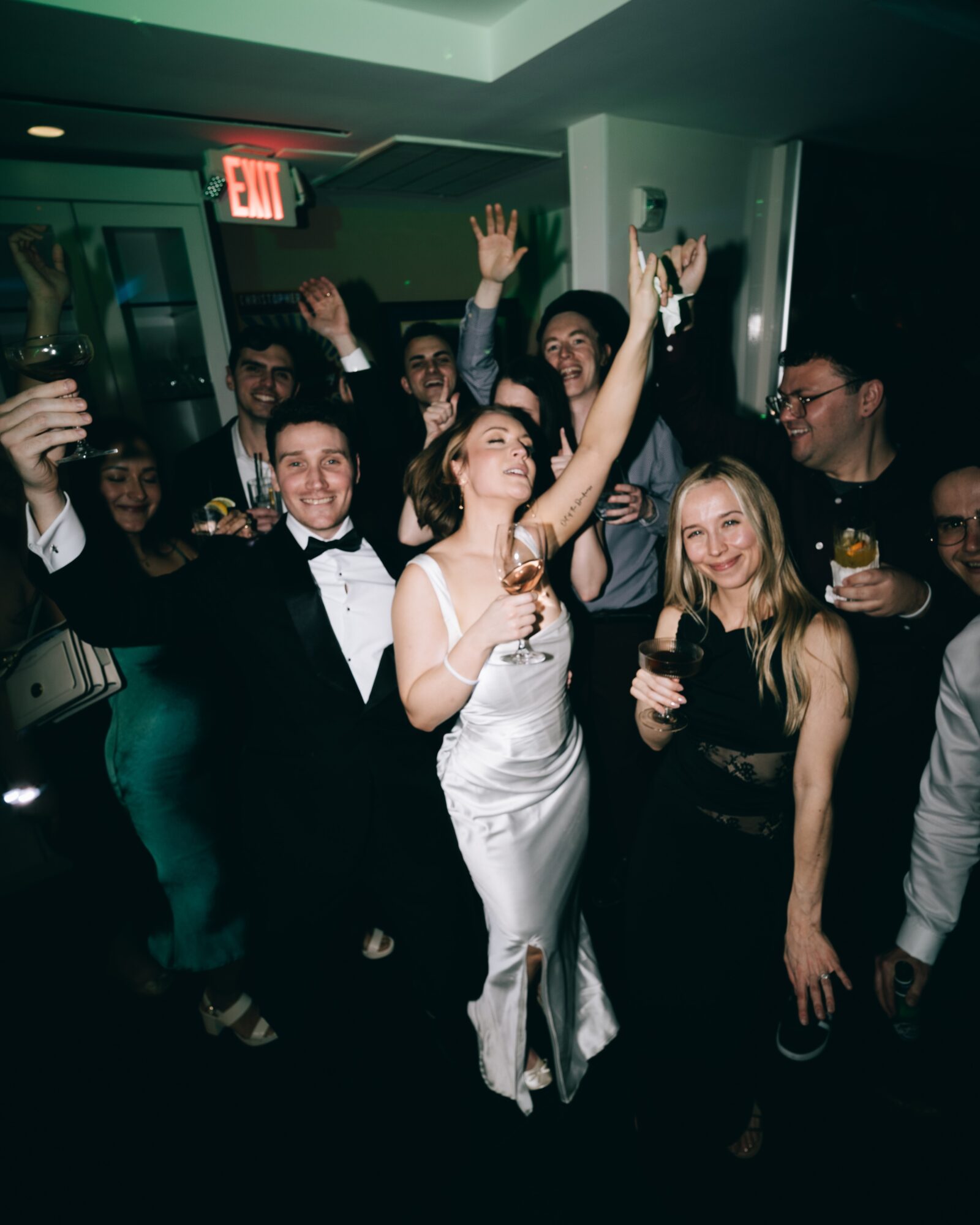 Group of people dressed in formal attire celebrating indoors, some holding drinks, with a woman in a white dress in the center.