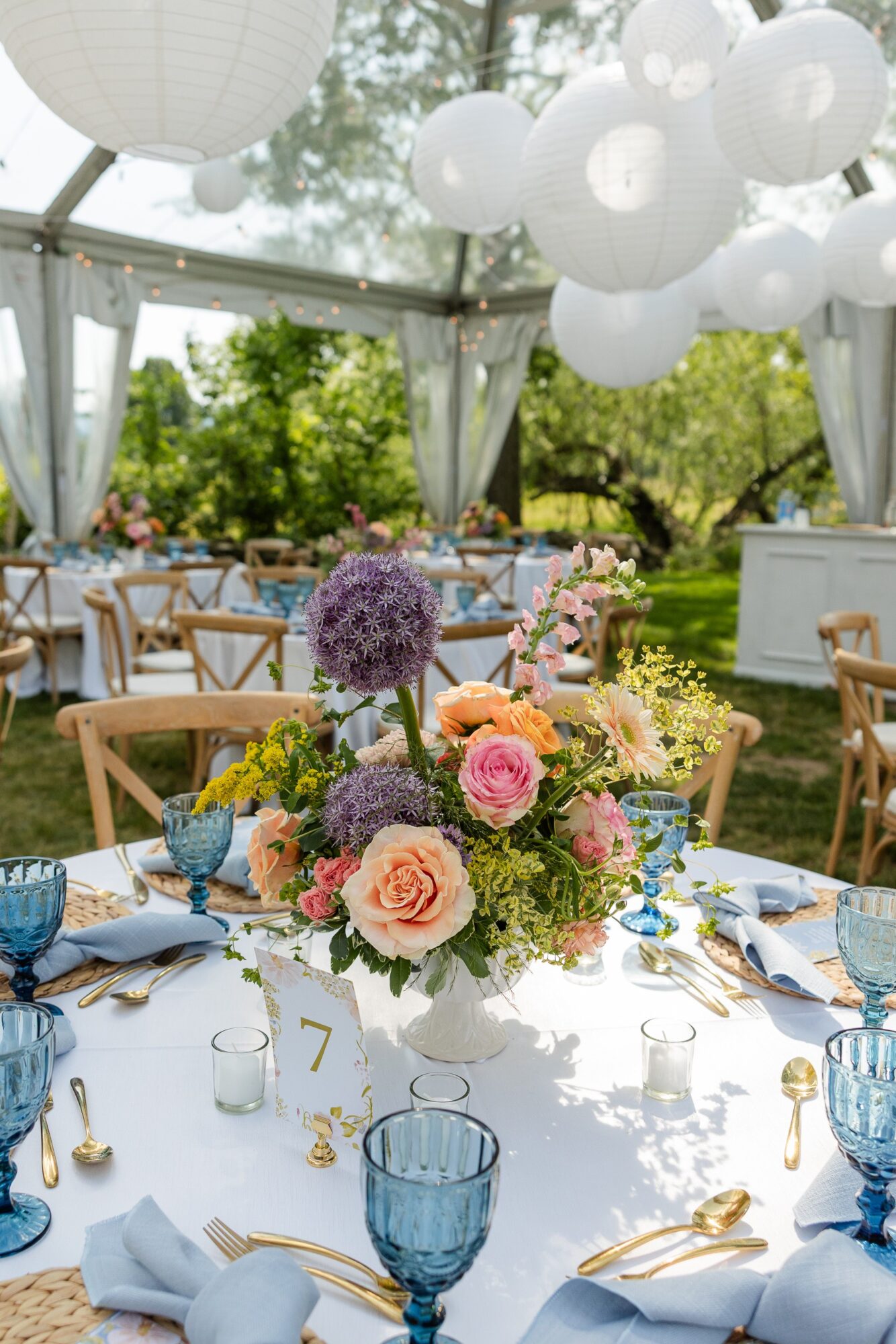 Elegant outdoor event setup with a floral centerpiece, tables, chairs, and hanging lanterns under a tent, surrounded by greenery.