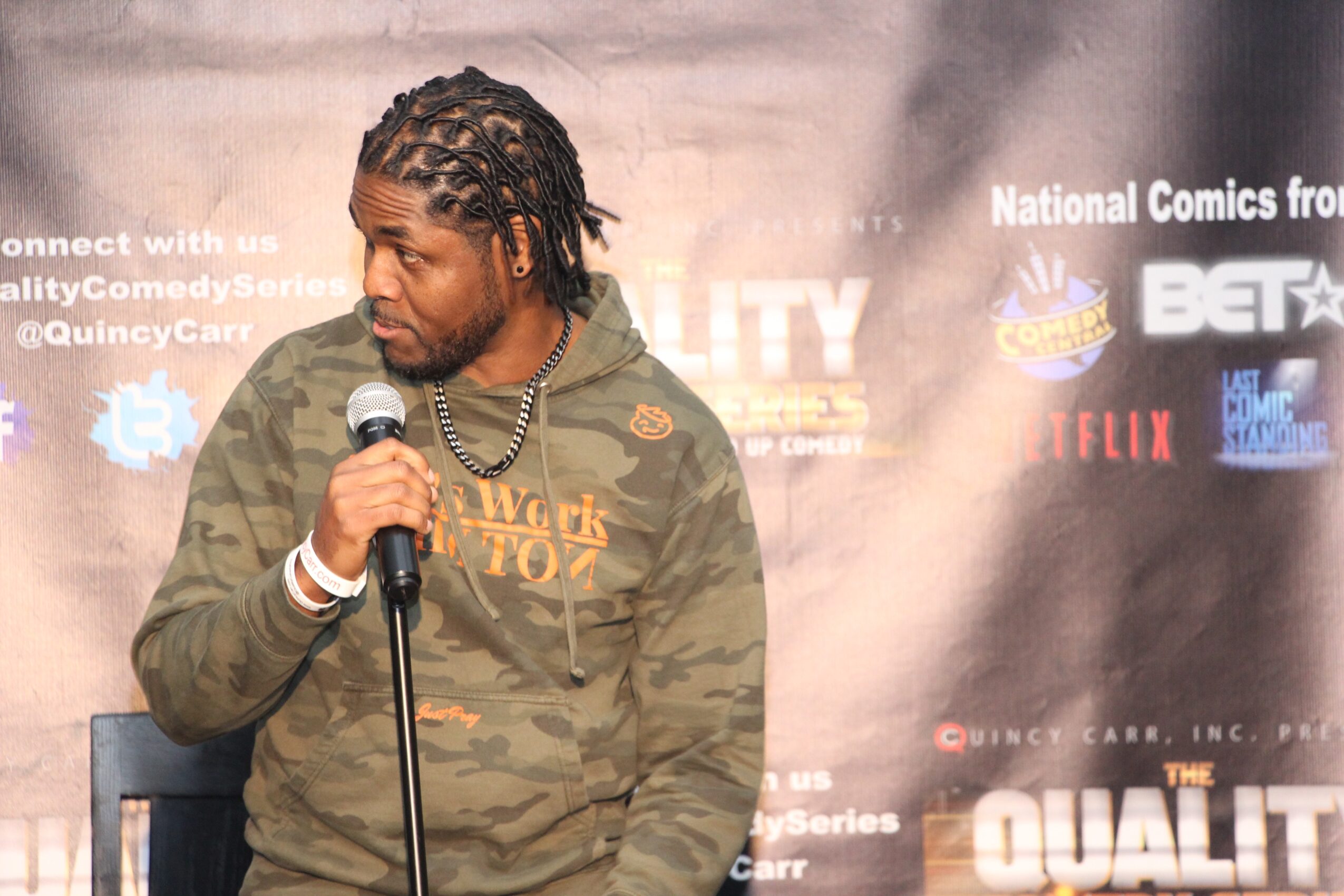 Man with dreadlocks holding a microphone, wearing a camouflage hoodie, standing in front of a promotional backdrop.