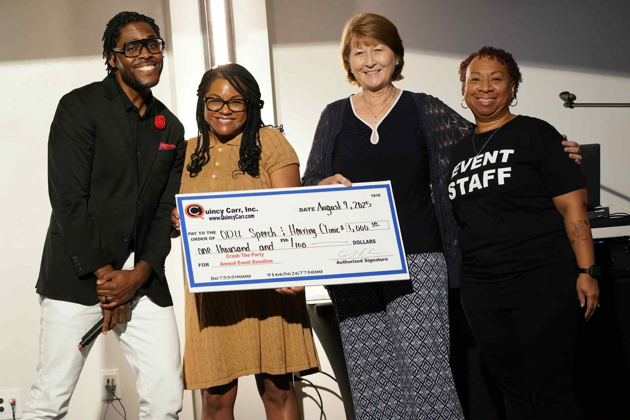 Four women standing together, one holding a large check, smiling at the camera, in an indoor setting.