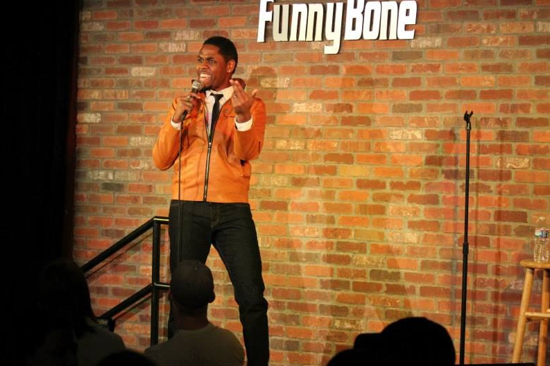 Stand-up comedian performing on stage with a brick wall background, holding a microphone, wearing an orange jacket and black pants.