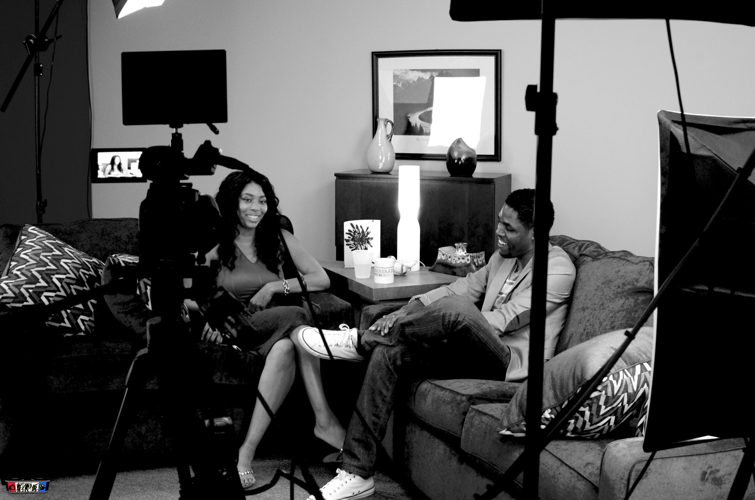 Two women sit on a sofa in a room with filming equipment, including lights and cameras, during an interview or recording session.