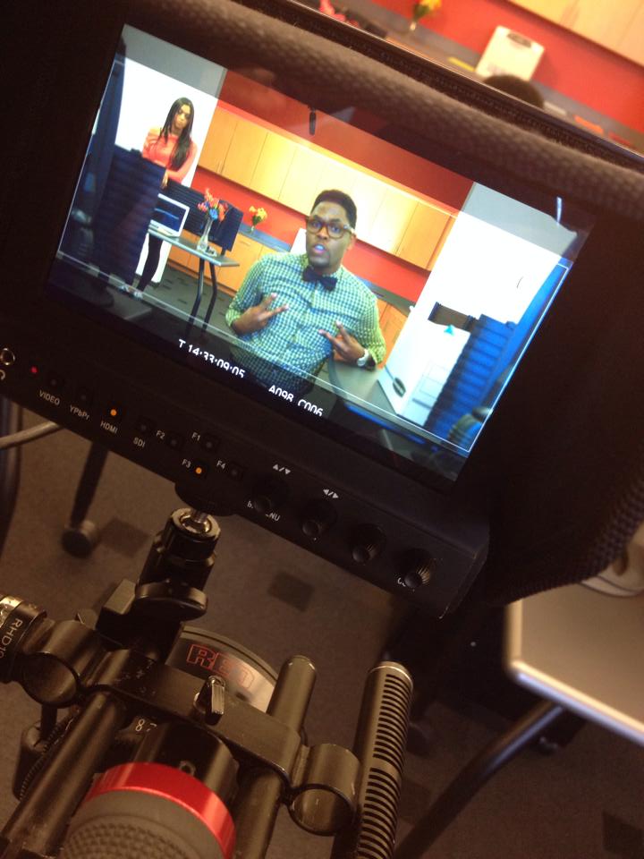 Television screen showing a man speaking in a room with colorful cabinets and a woman in the background.