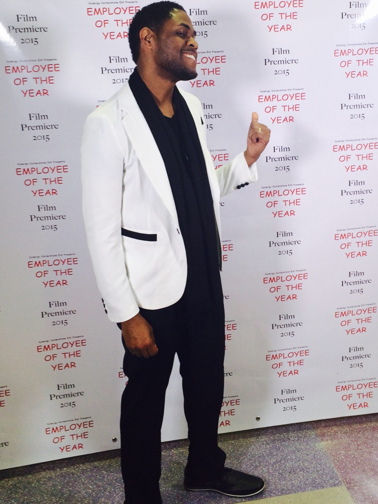 Man in white blazer and black pants standing in front of a backdrop with red text, smiling and gesturing with his right hand.