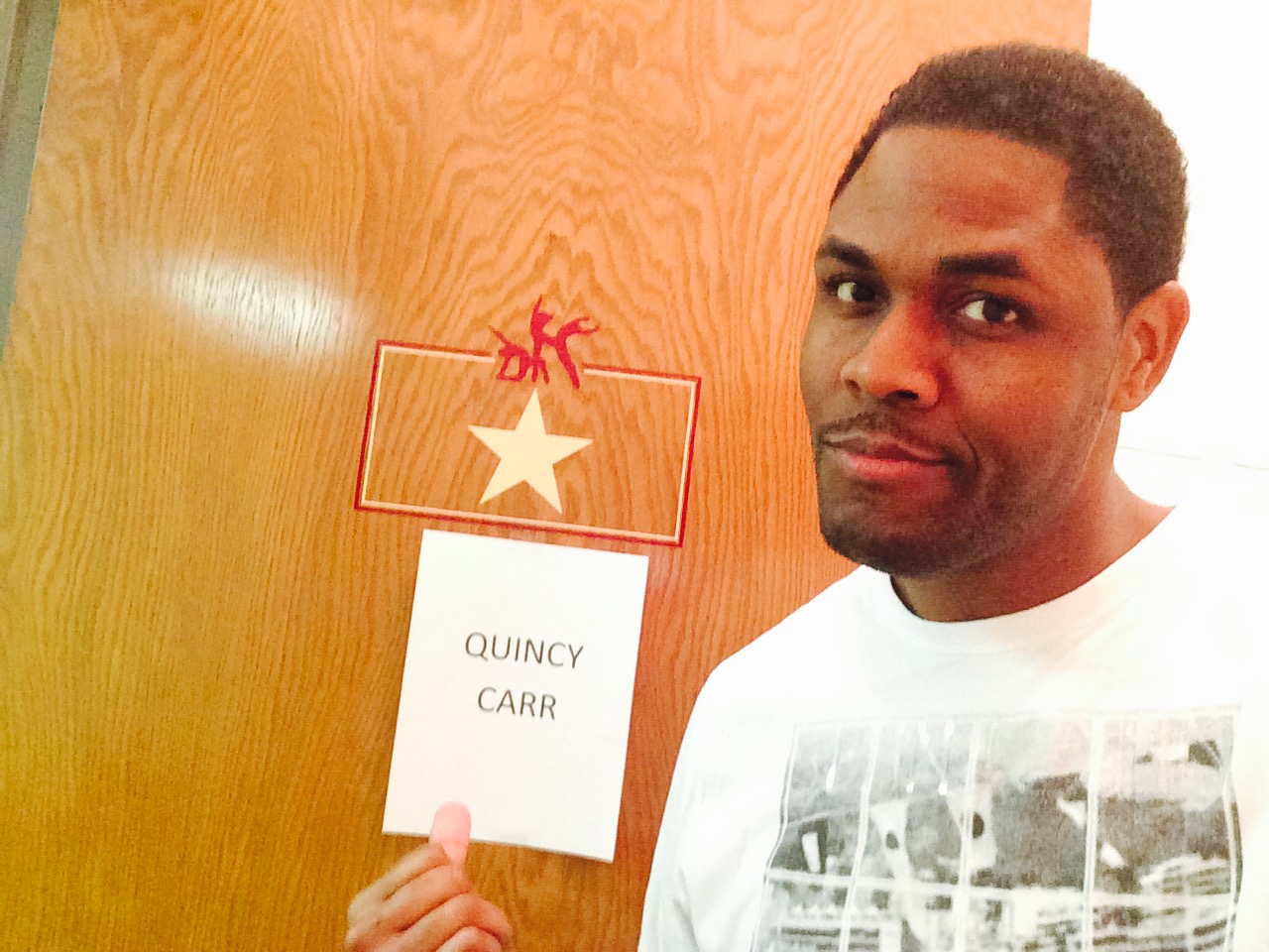 Man with short hair and white shirt standing next to a door with a name tag reading 'QUINCY CARR' and a star decoration.