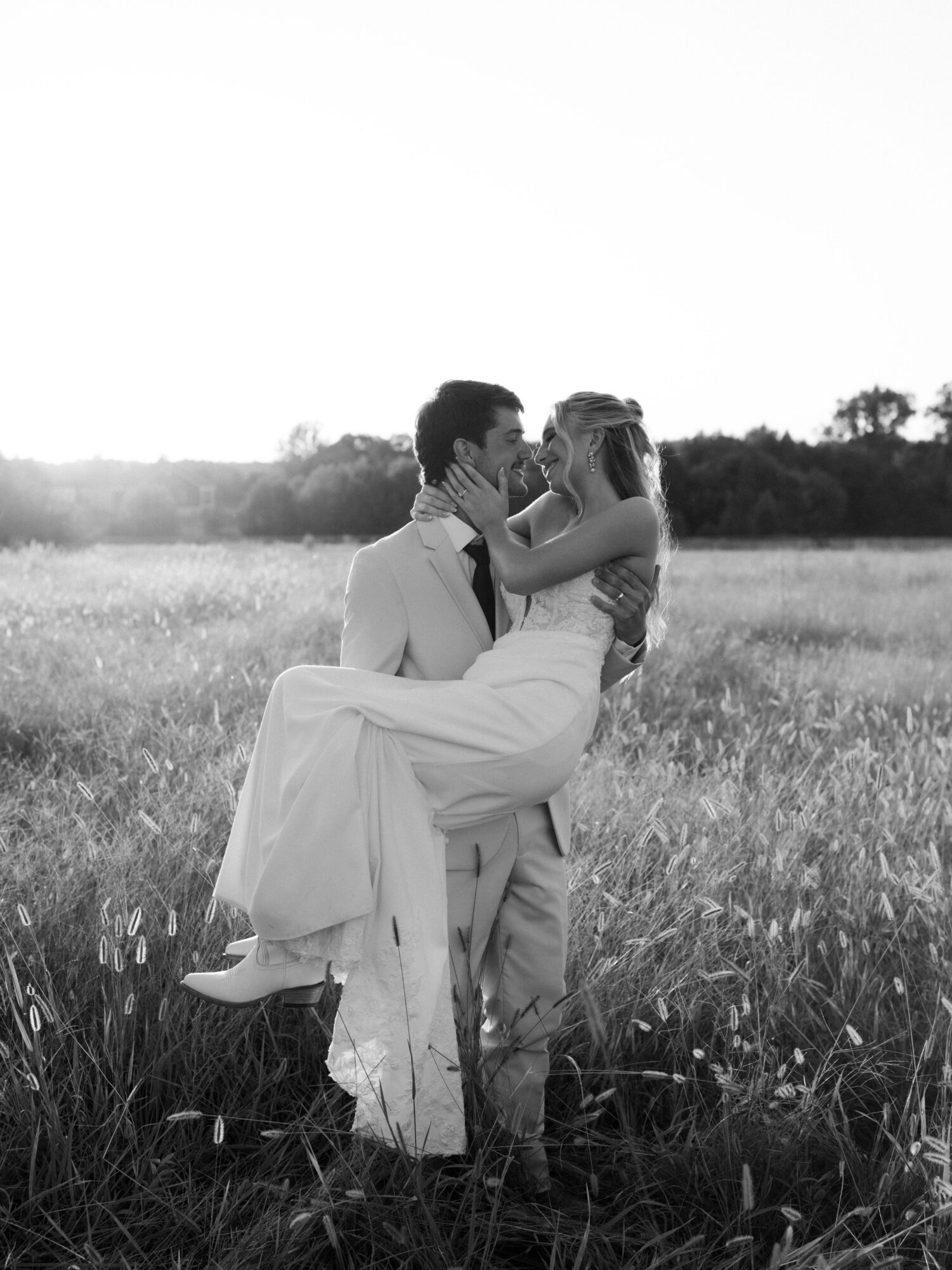 A couple in wedding attire embracing in a field, with the man lifting the woman, both smiling, in black and white.