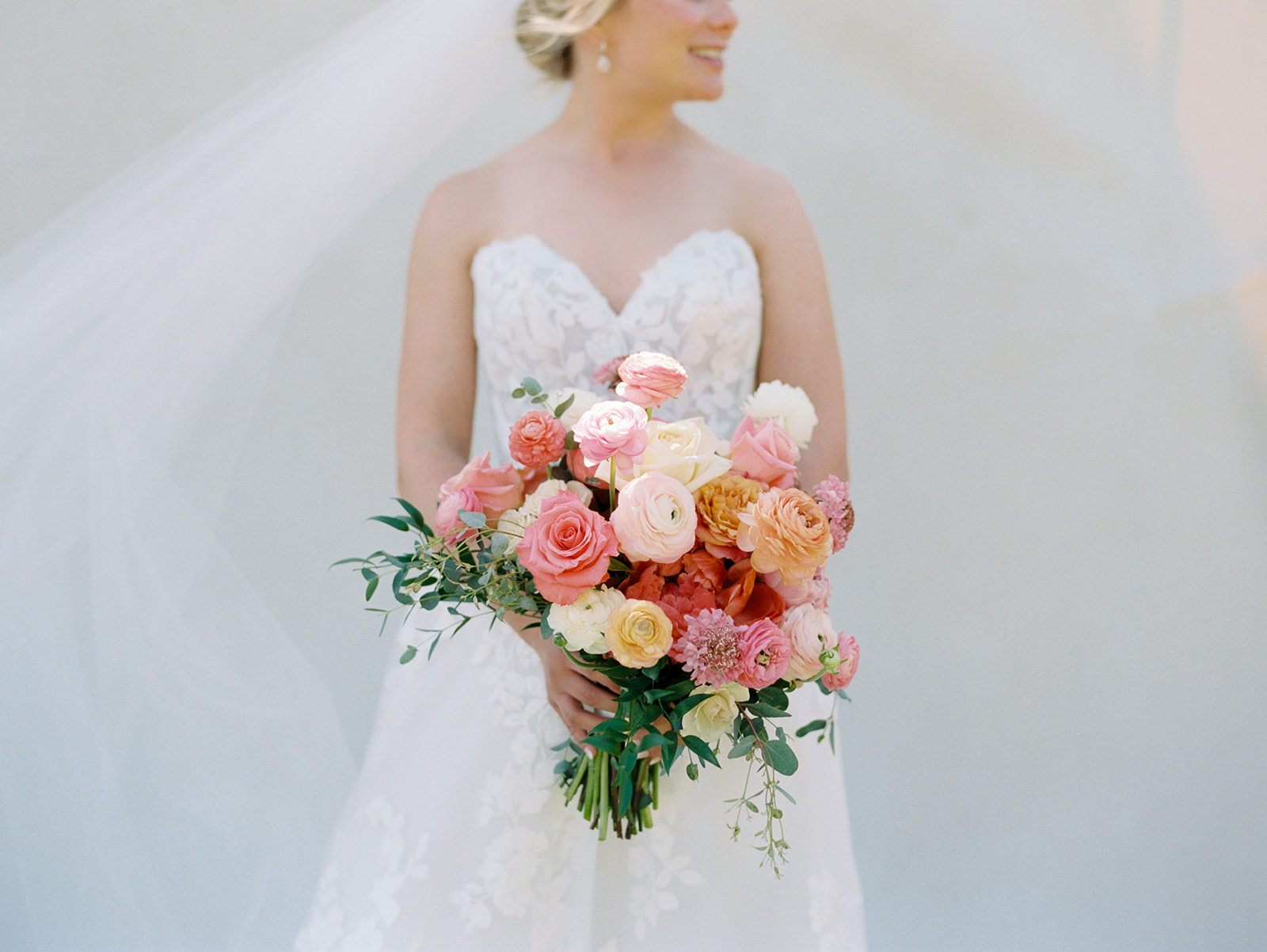 Bride in a white wedding dress holding a large bouquet of pink, peach, and cream flowers, smiling, veil flowing behind her.