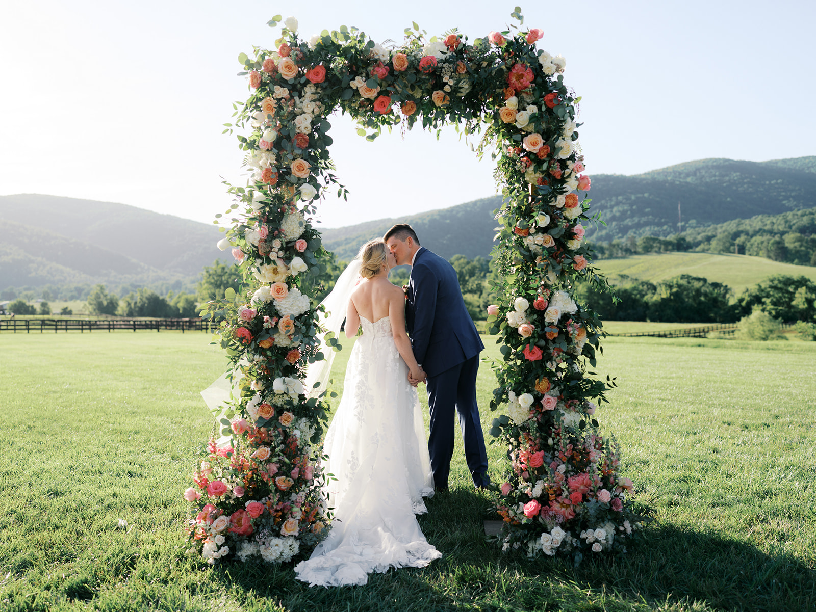 Bride and groom kissing under a large floral arch in a green field with hills in the background.
