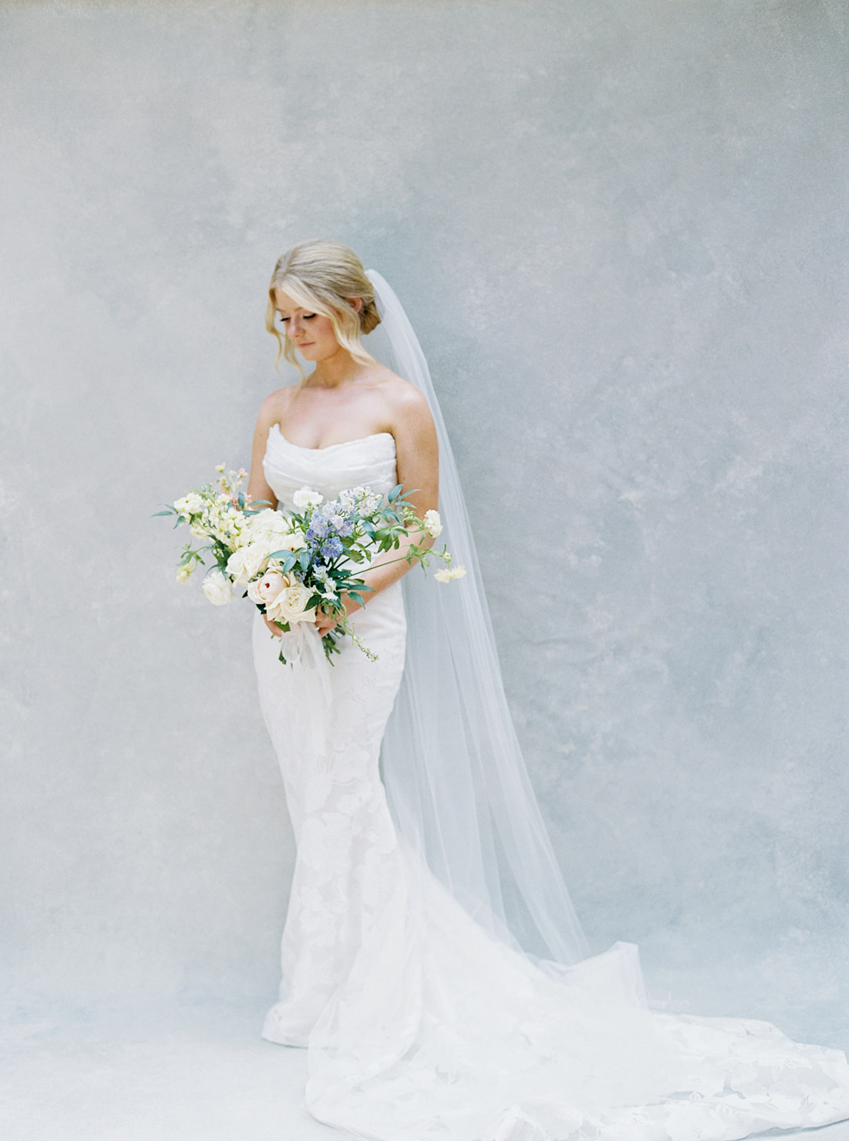 Bride in a white wedding dress holding a bouquet of flowers, standing against a plain light gray background.