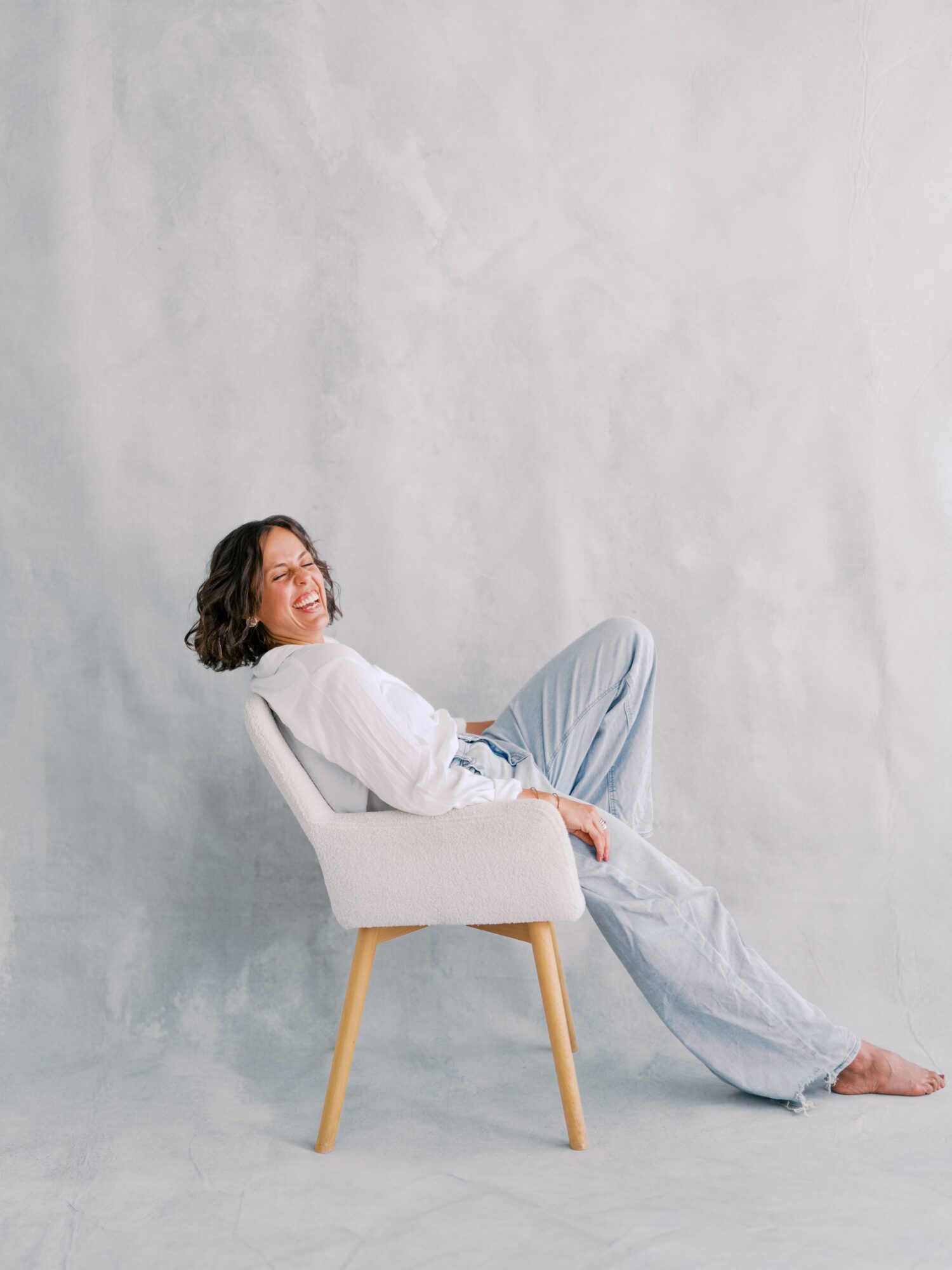Woman sitting on a white chair, smiling, with a plain light gray background.