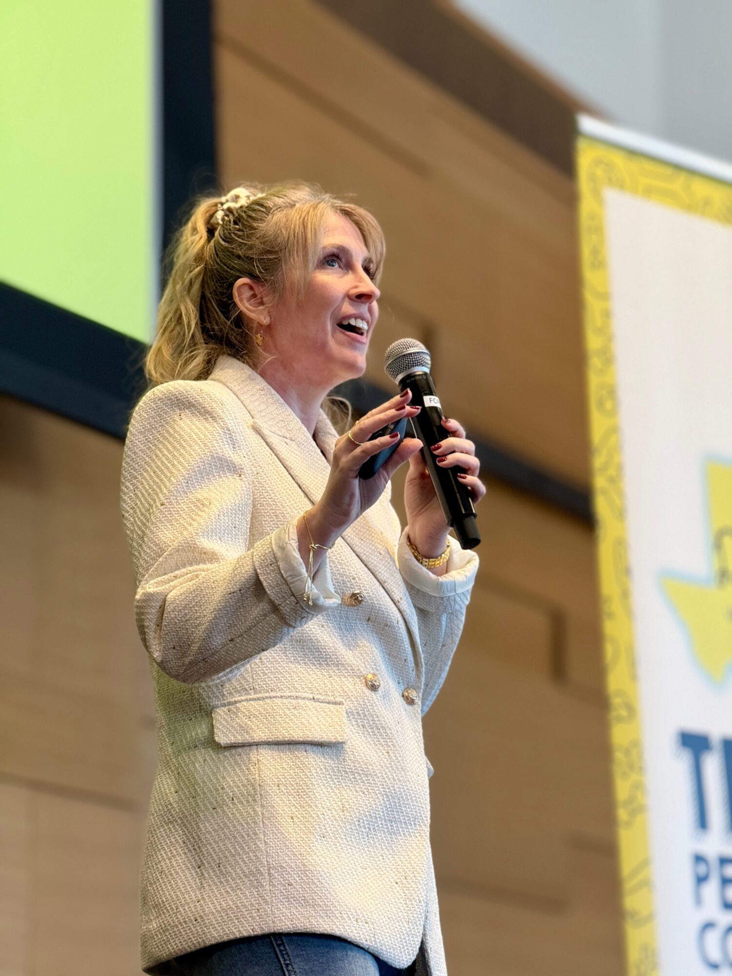 Woman speaking into a microphone, smiling, wearing a light-colored blazer, in front of a yellow and white banner.