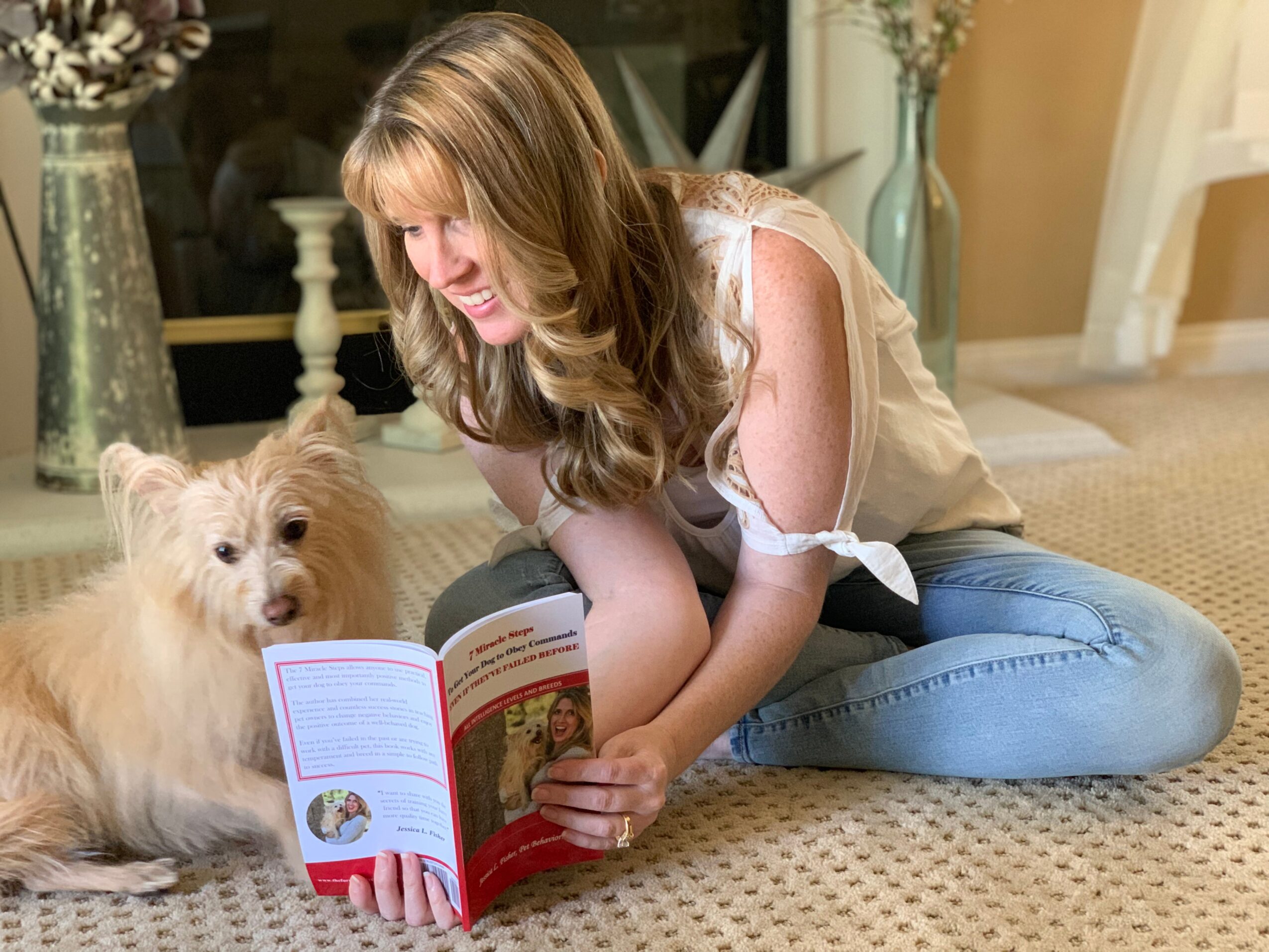Woman sitting on carpet reading a book with a small dog nearby, in a cozy room with curtains and furniture.