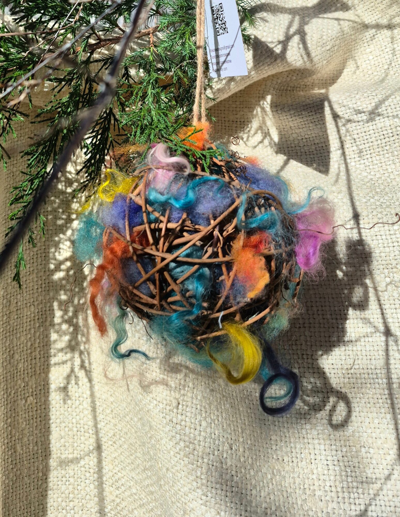 Colorful handmade bird ornament with feathers, twigs, and string hanging from a tree branch, casting a shadow on a textured surface.