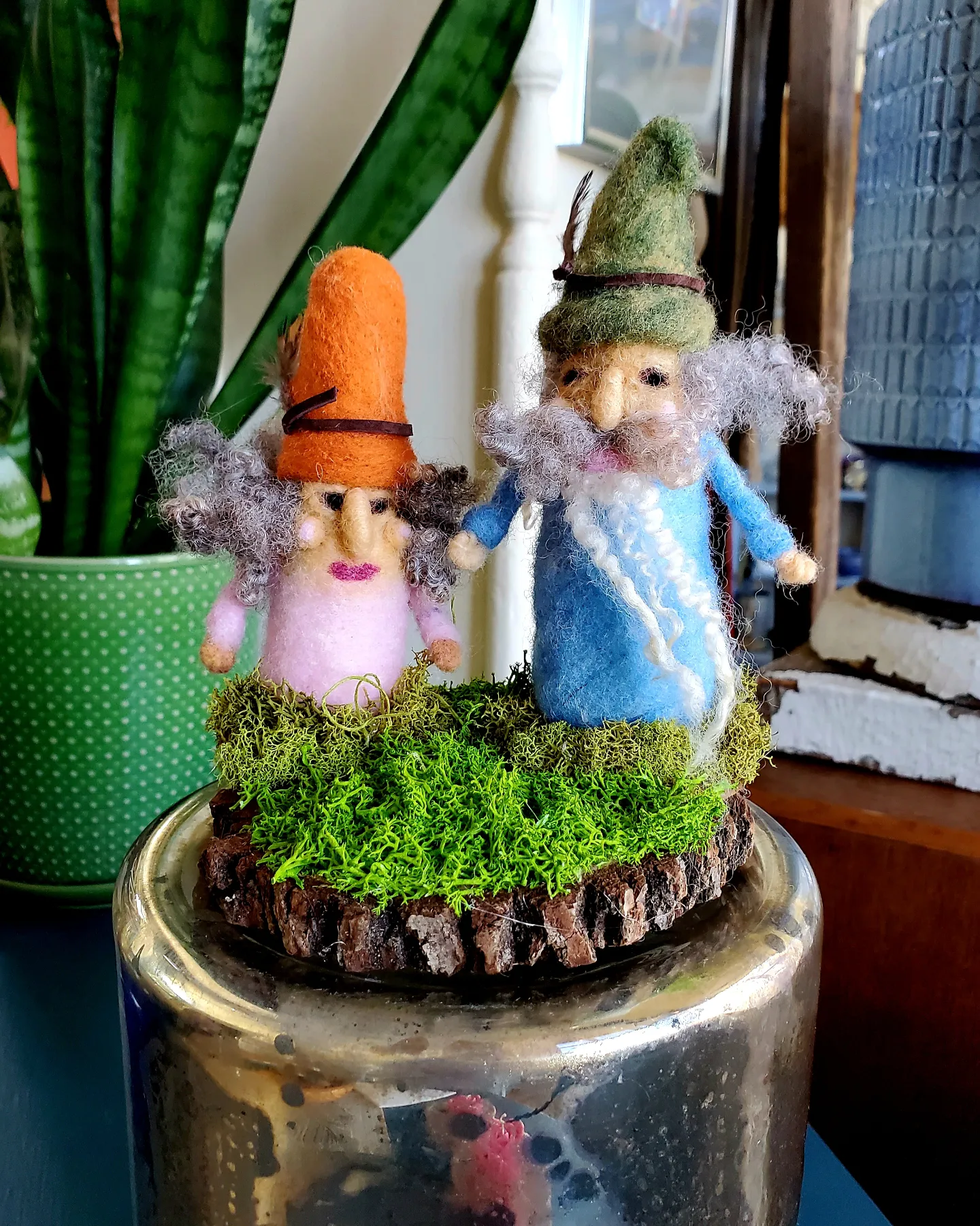 Two felted gnome figures with hats stand on a moss-covered wooden base, placed on a metallic surface indoors.