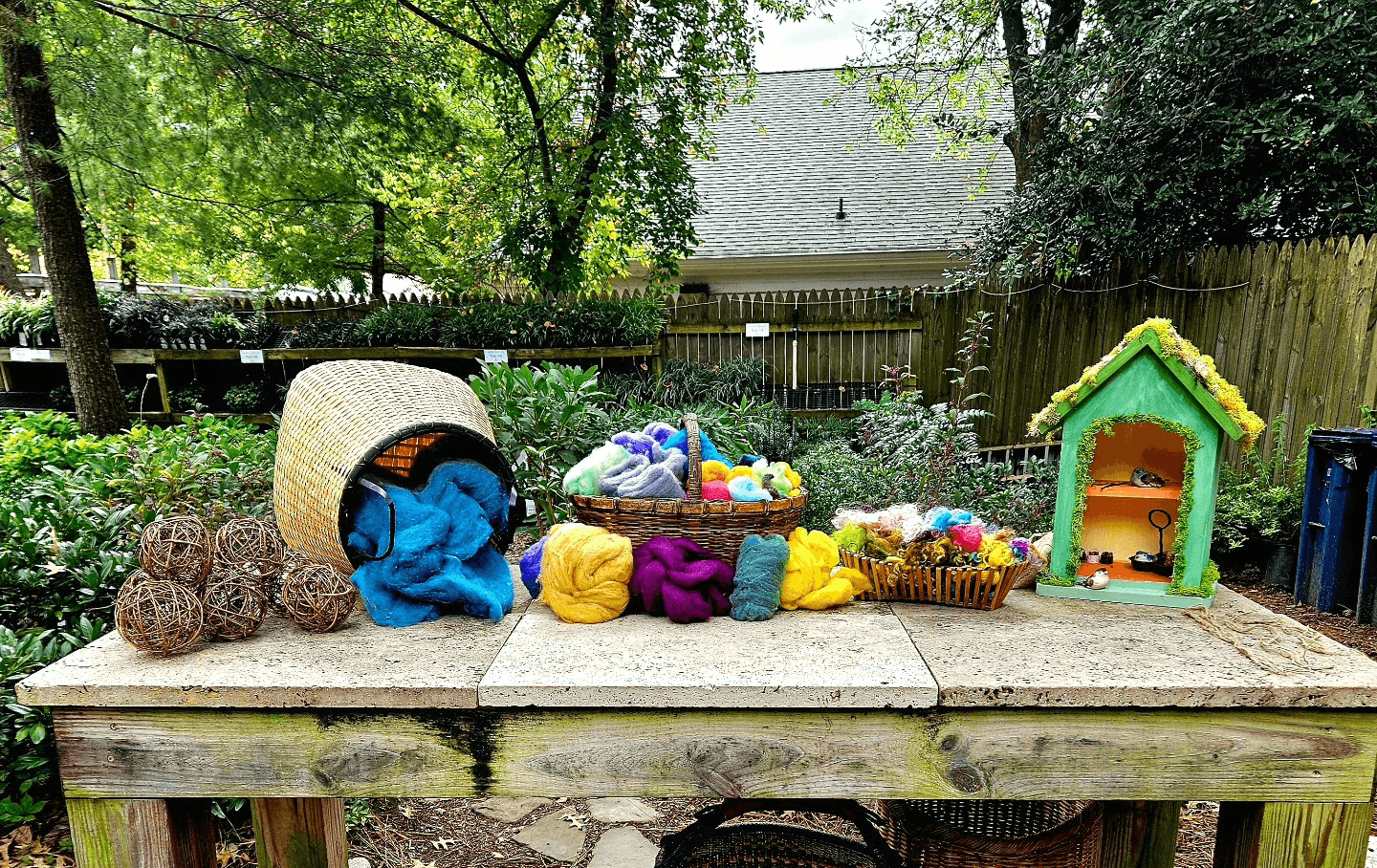 Outdoor scene with toys, baskets, and a small playhouse on a wooden table, surrounded by trees and a fence.