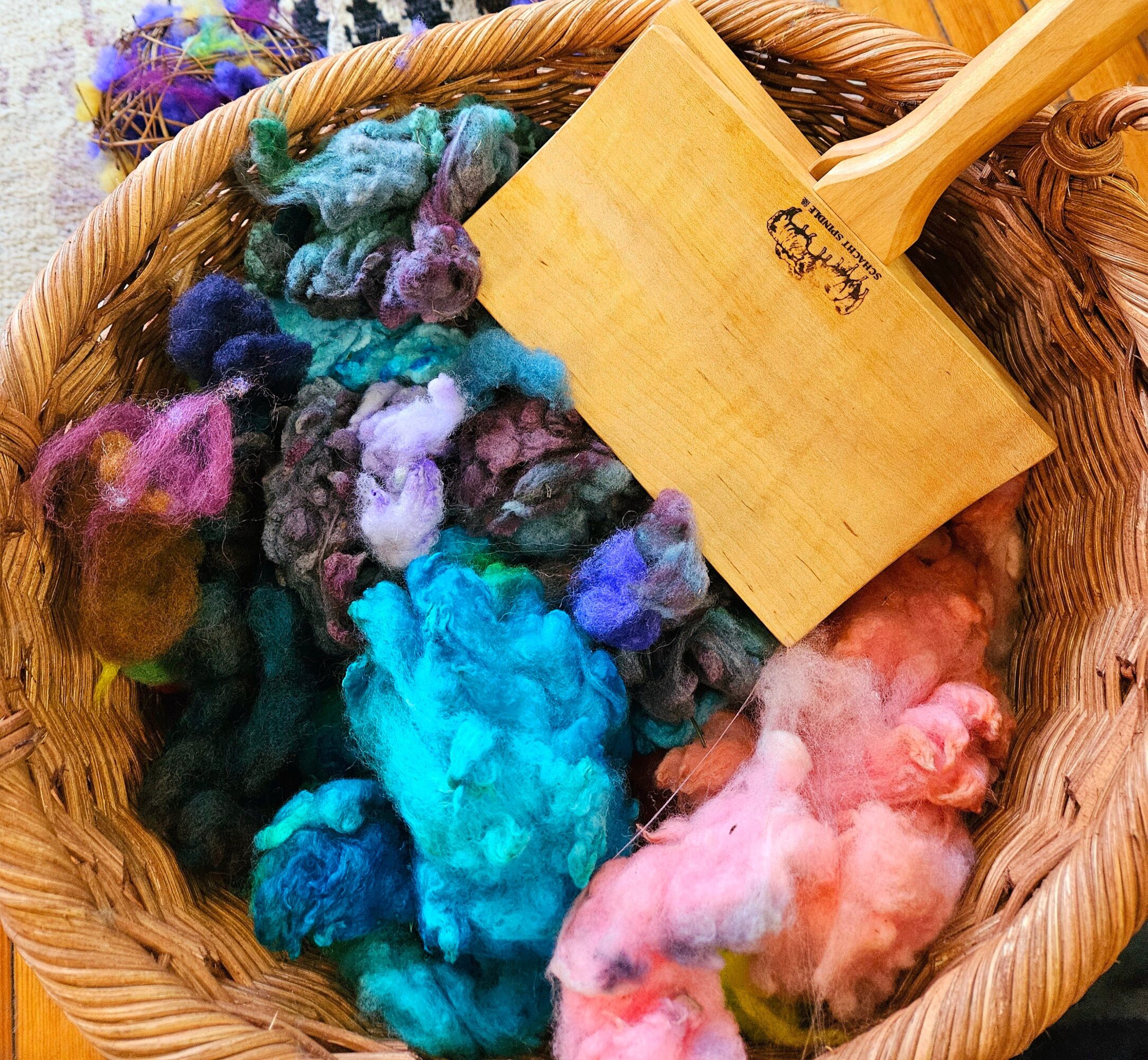Colorful wool fibers in a basket with a wooden tool, various shades including blue, pink, purple, and green.