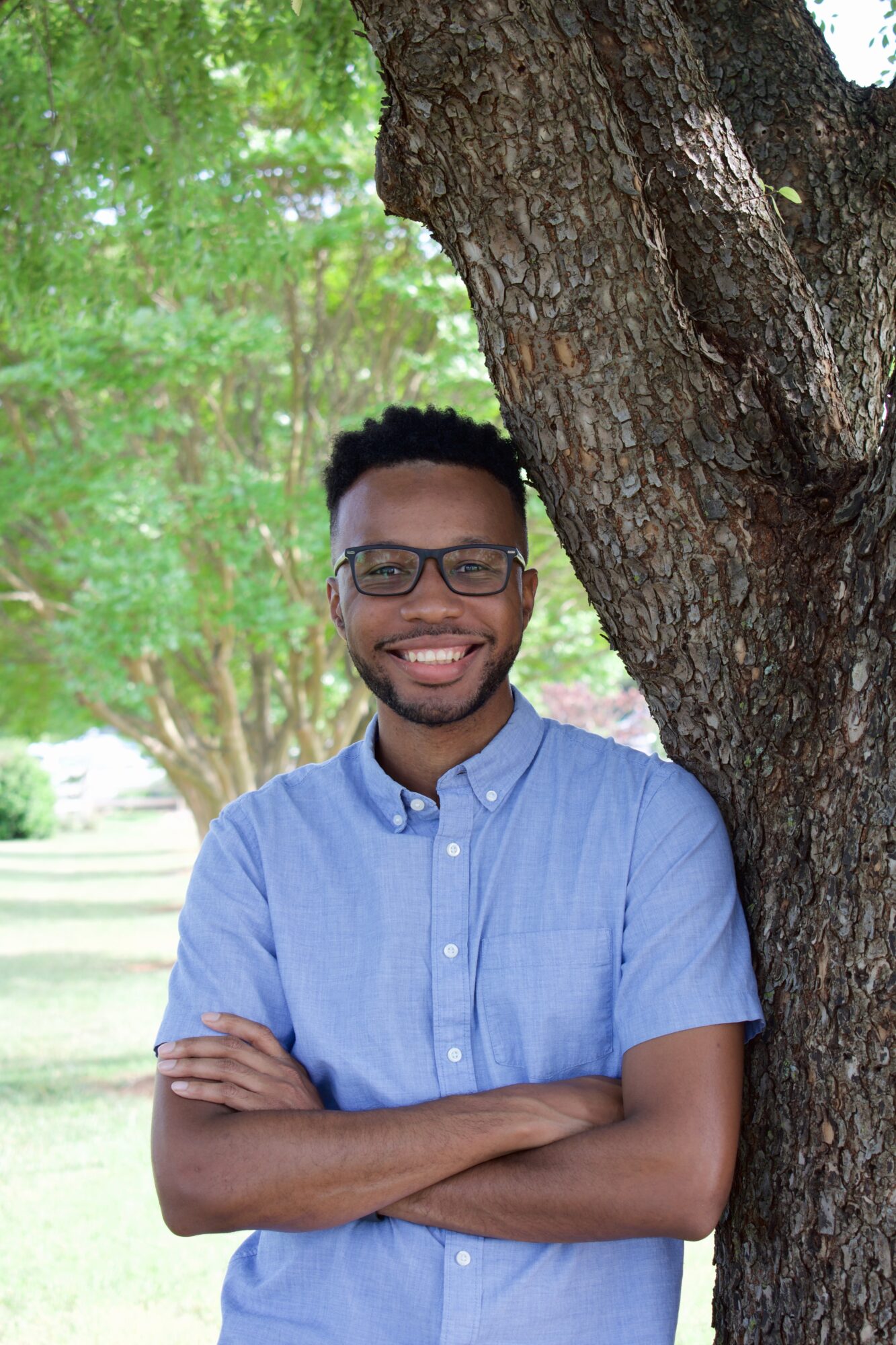 Young man with glasses smiling outdoors, standing next to a tree with arms crossed, wearing a light blue shirt.