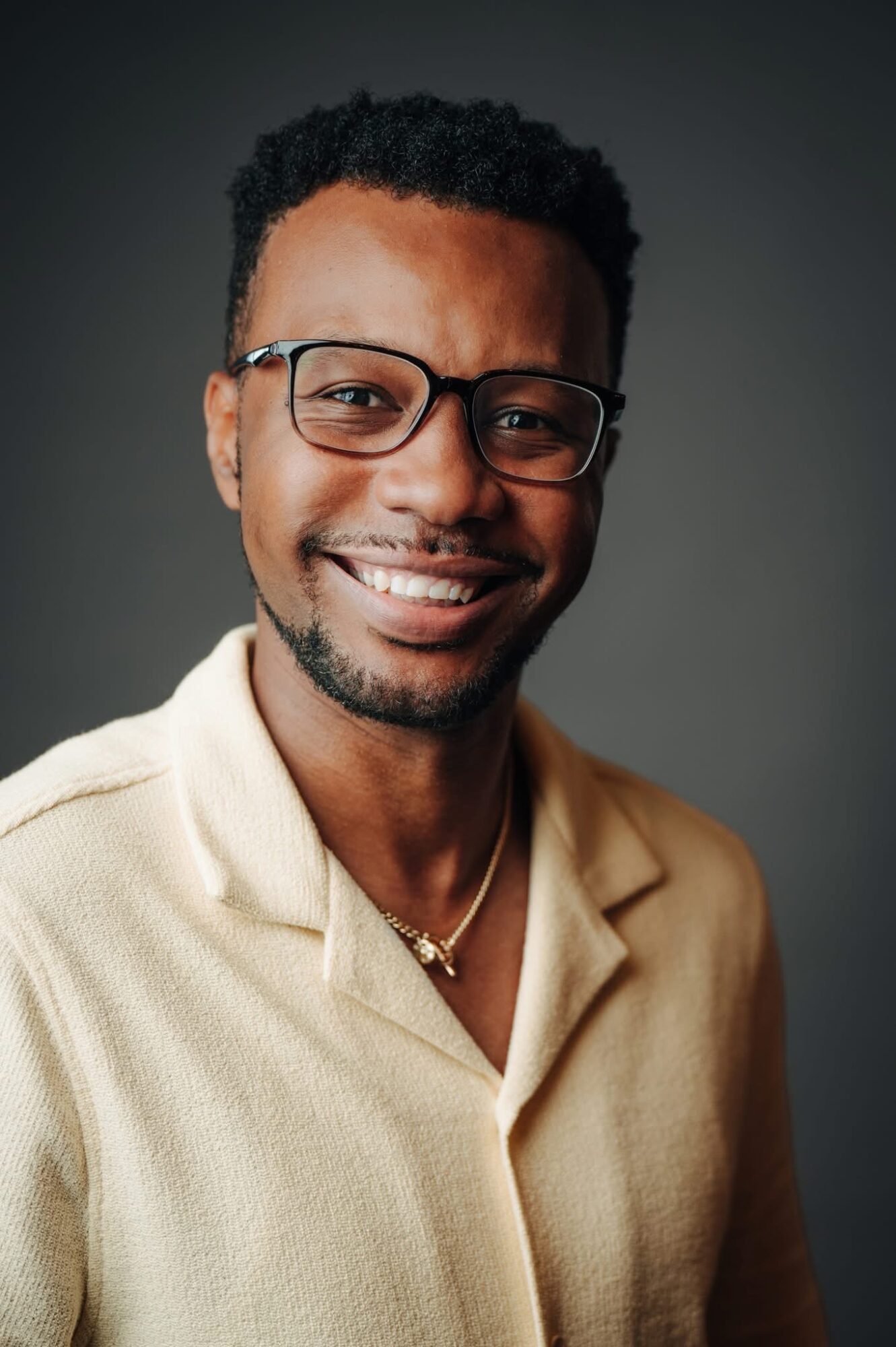 Smiling man with glasses and short curly hair, wearing a beige shirt and necklace, against a dark background.