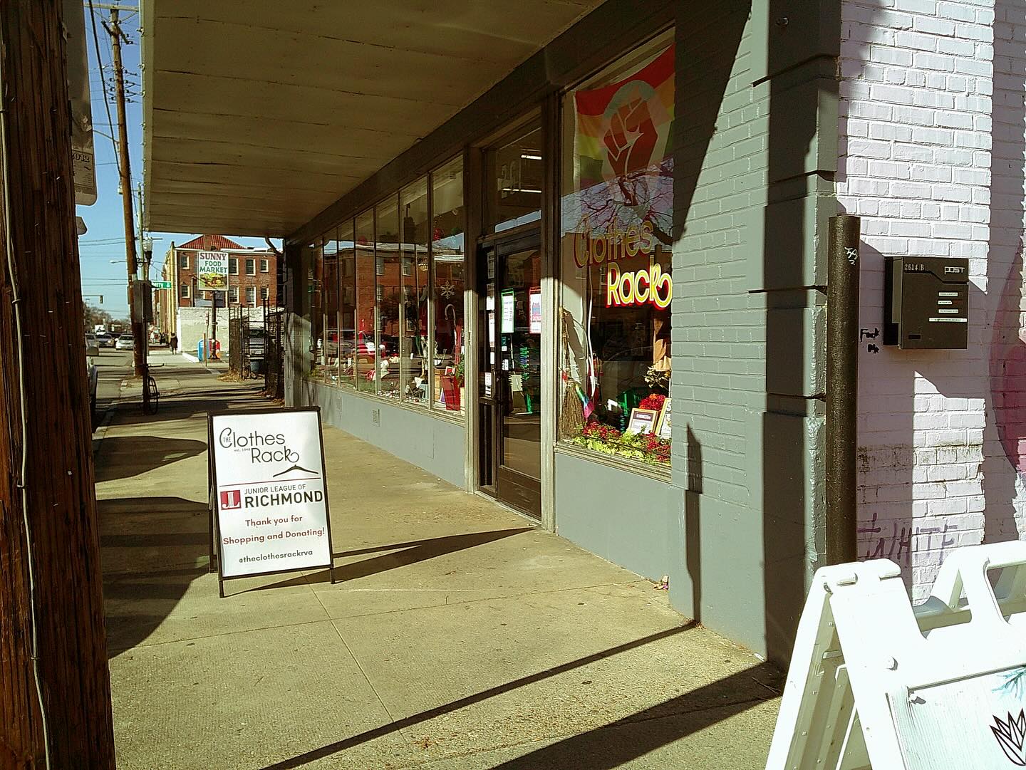 Storefront with large glass windows and a sidewalk sign outside, sunlight casting shadows, brick building on the right.