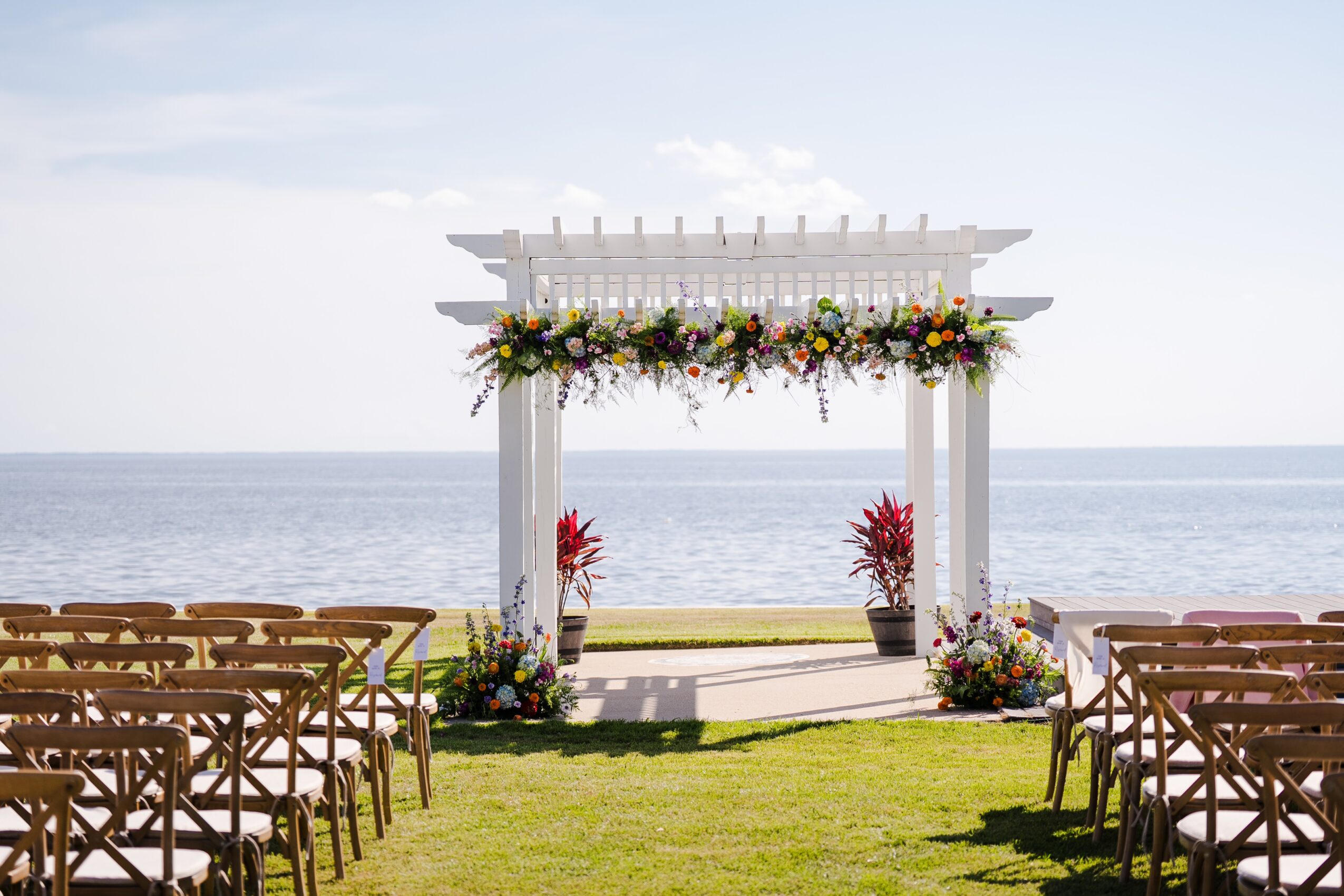 Wedding arch decorated with flowers on a grassy area by the water, with chairs arranged on both sides.