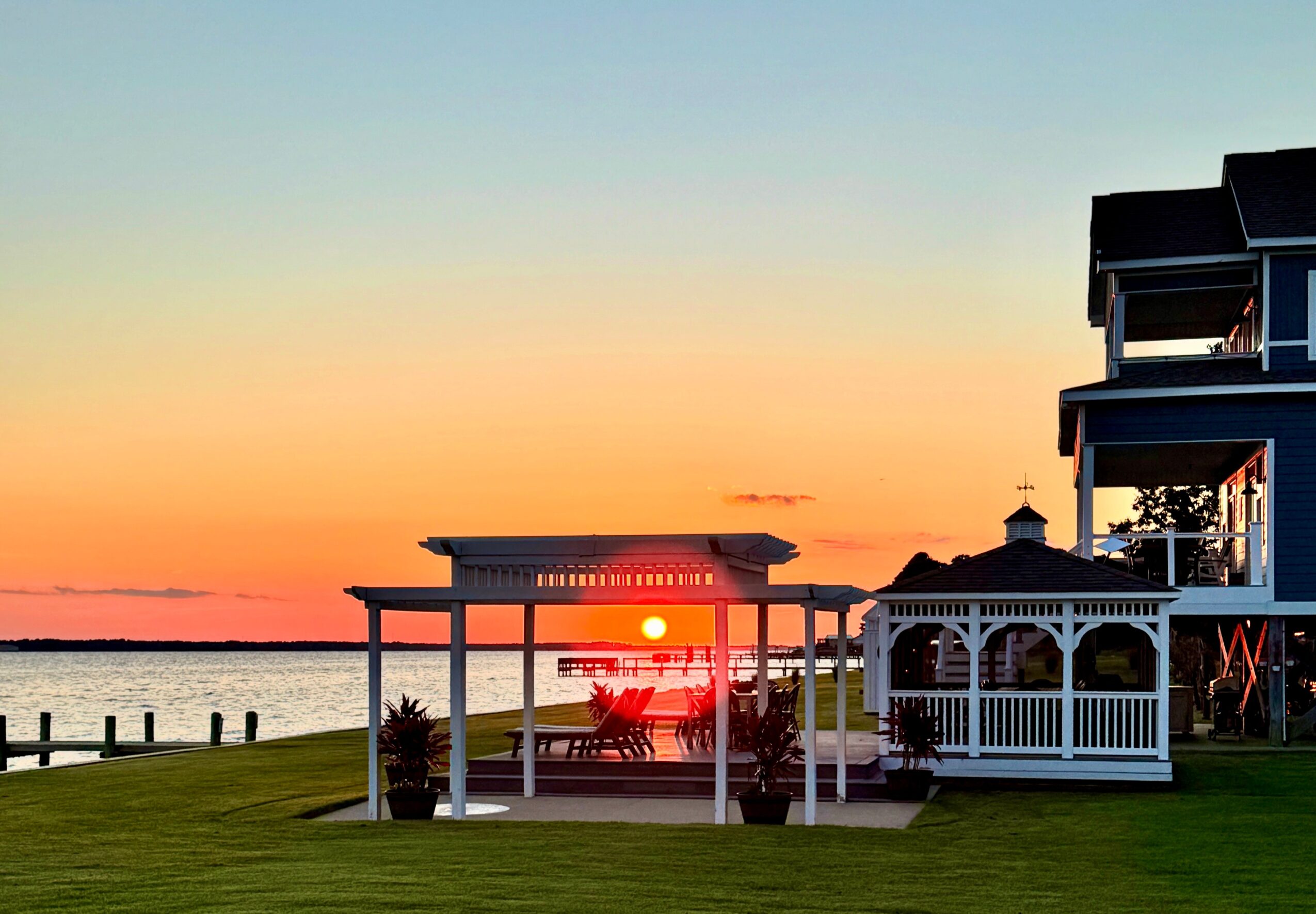 Sunset over water with a pavilion and a building nearby, colorful sky, grassy area in foreground.