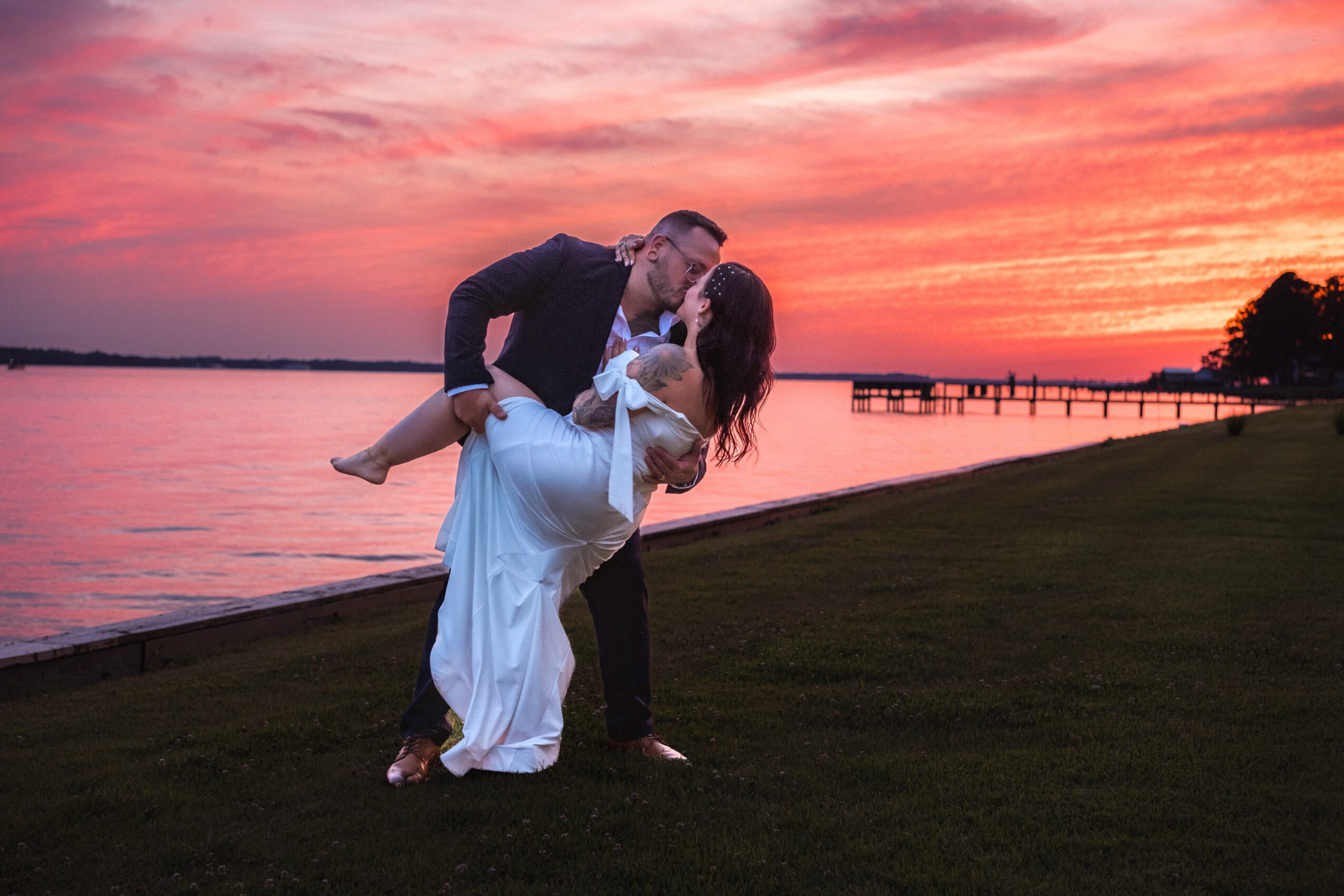 Couple kissing outdoors at sunset near water, with a pier in the background, grassy area in foreground.