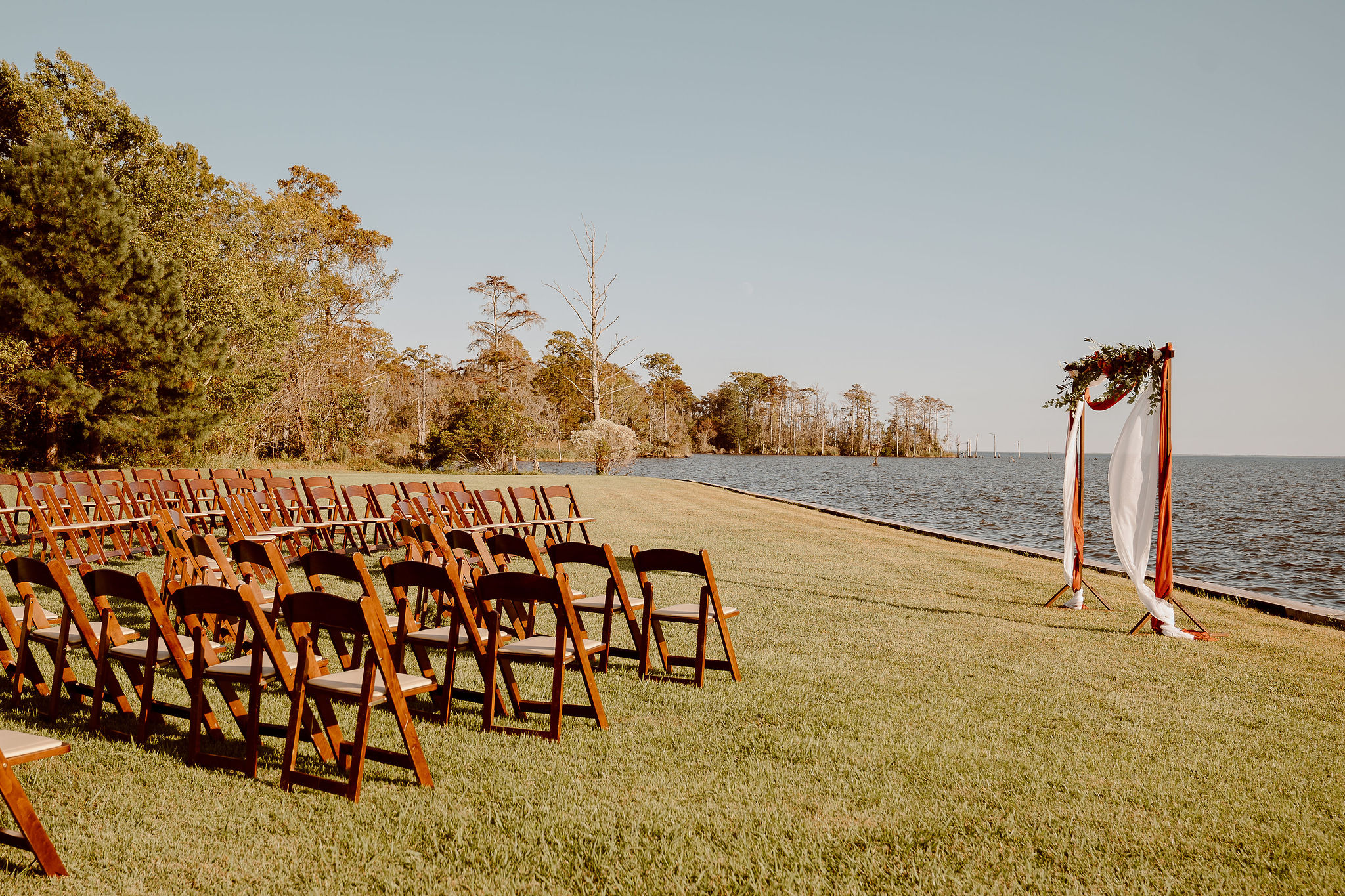 Outdoor wedding setup with chairs facing a decorated arch by the water, grassy area, and trees in the background.