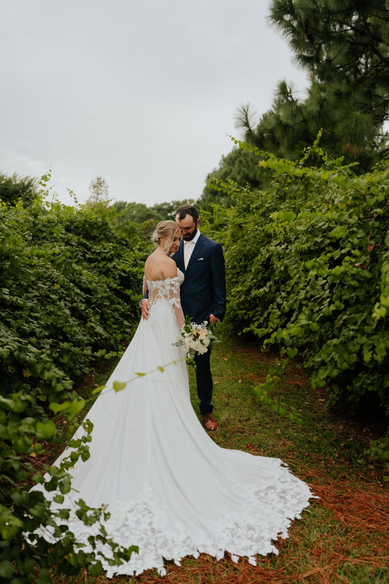 Bride and groom stand close together on a garden path surrounded by green bushes and trees, bride holding a bouquet.