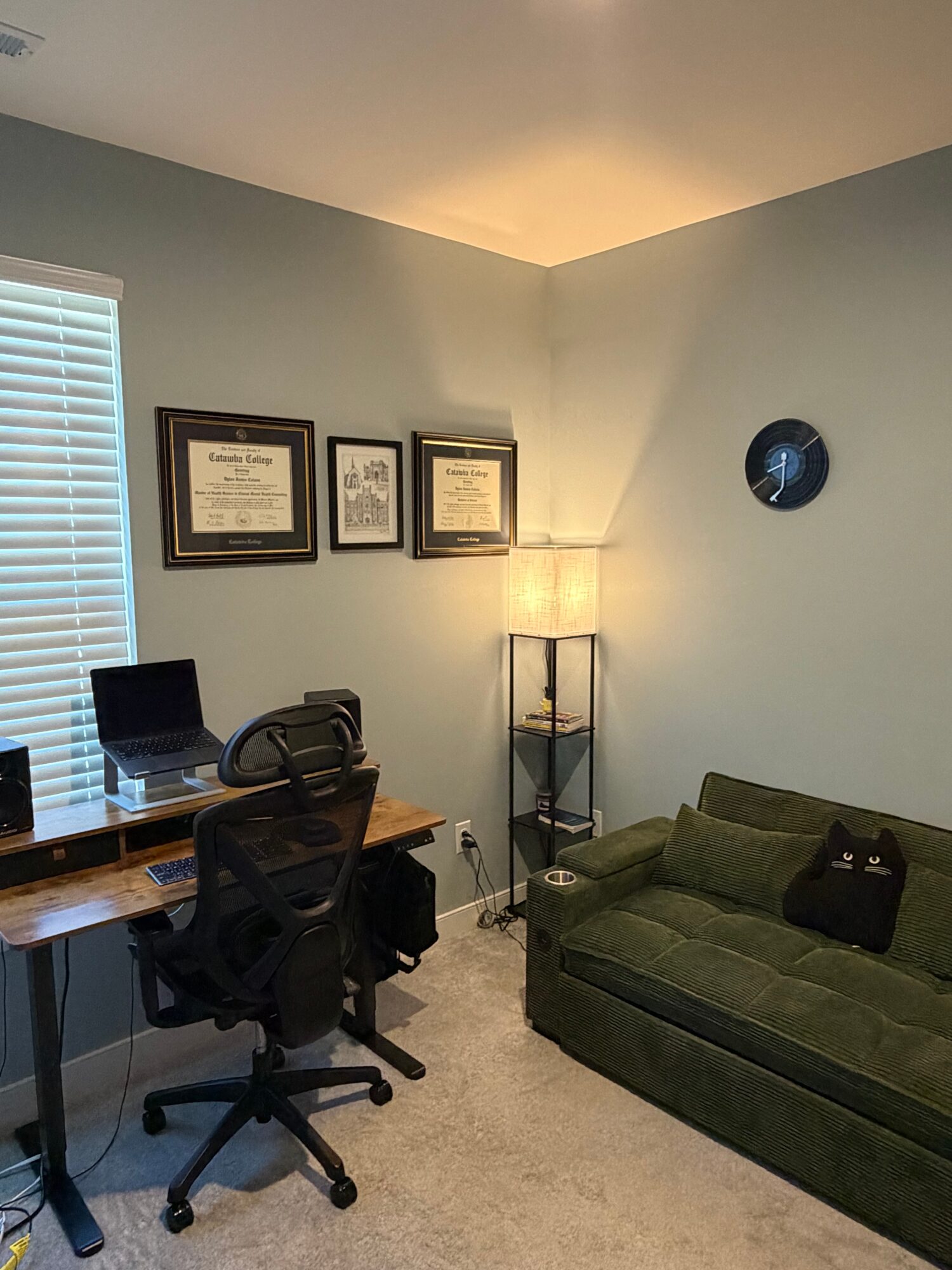 Corner of a room with a desk, chair, lamp, green sofa, and framed certificates on the wall.