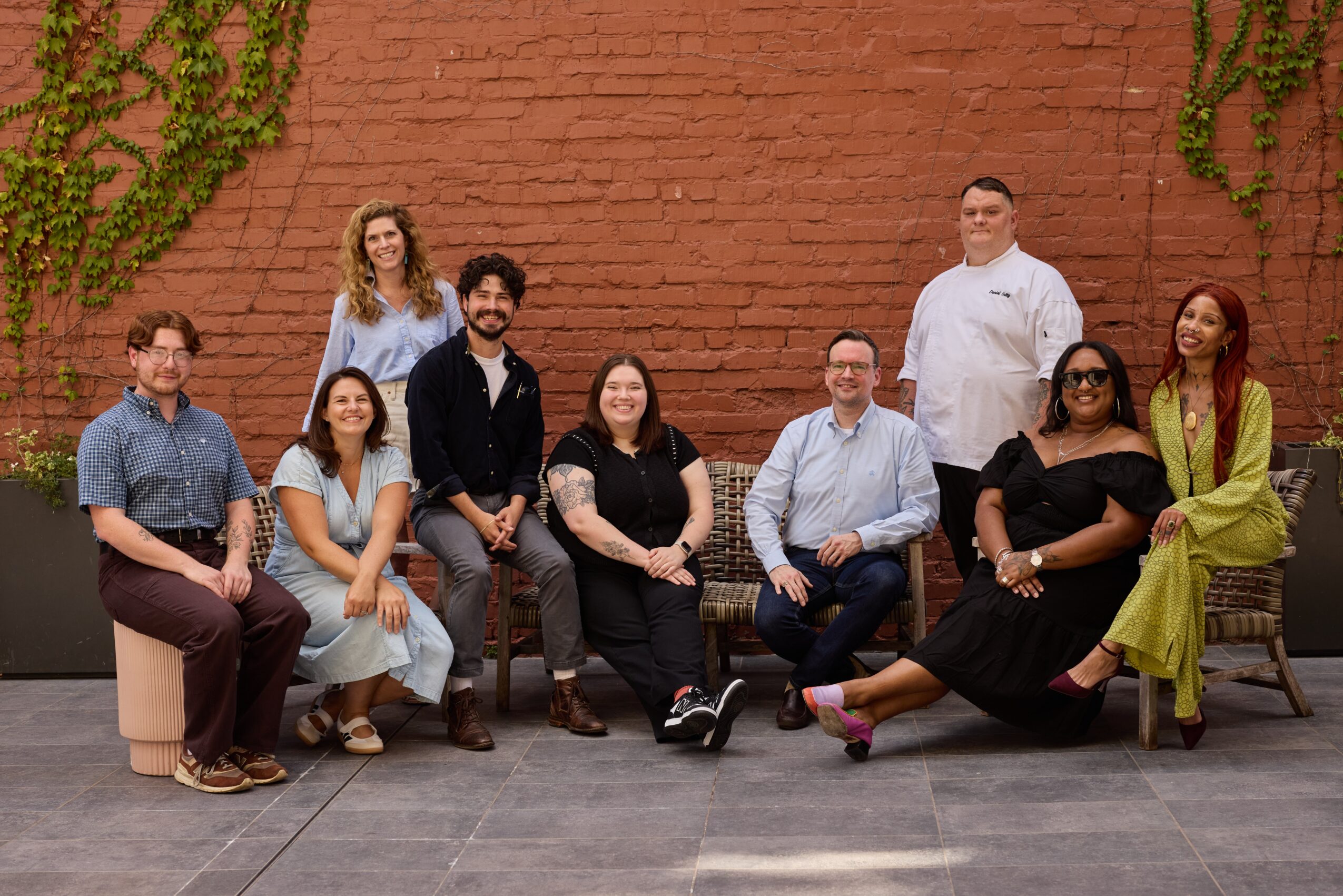 Group of nine people sitting and standing against a brick wall with green vines, smiling and posing for a photo.