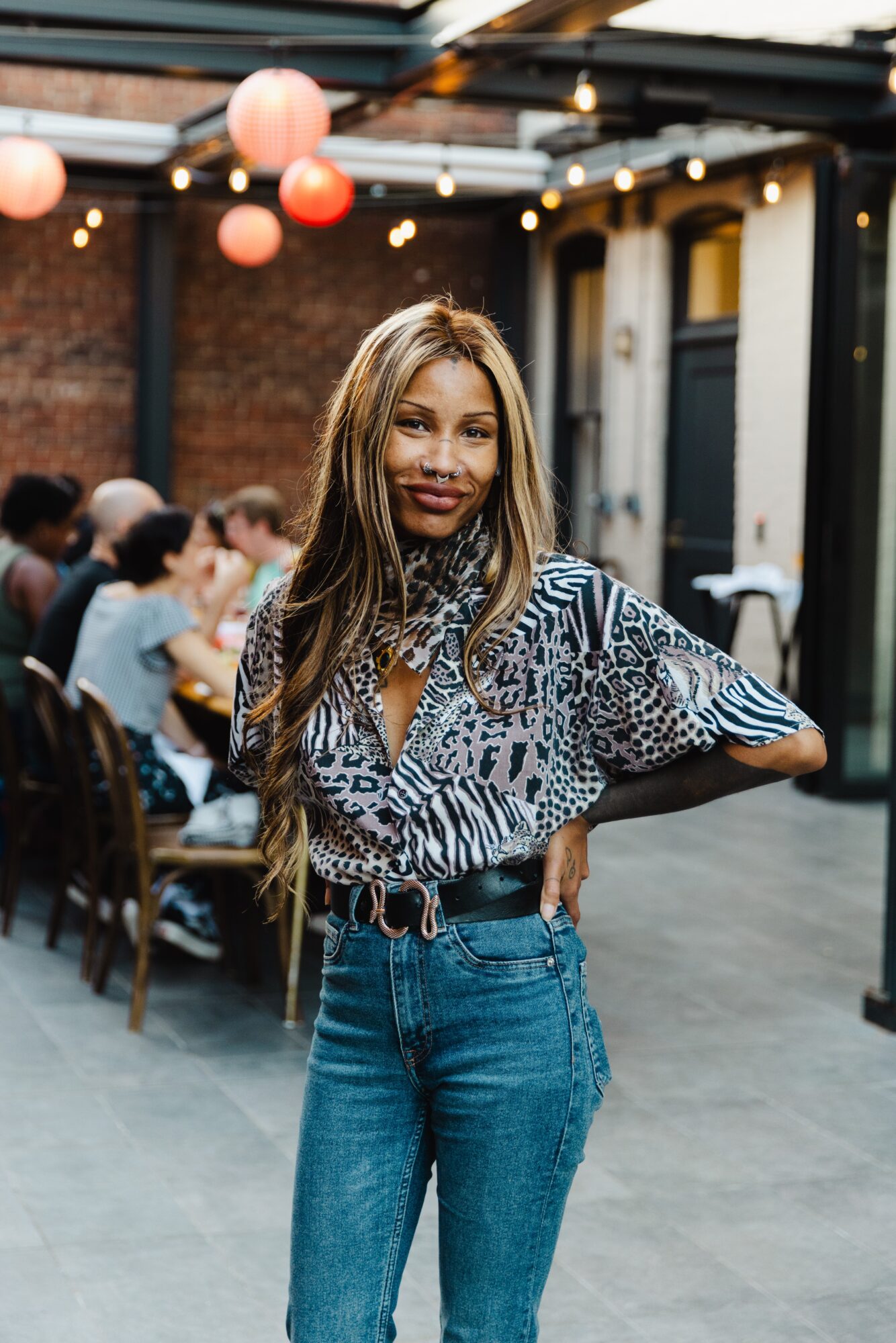 Woman with long hair wearing a patterned blouse and jeans, standing outdoors with people dining in the background.