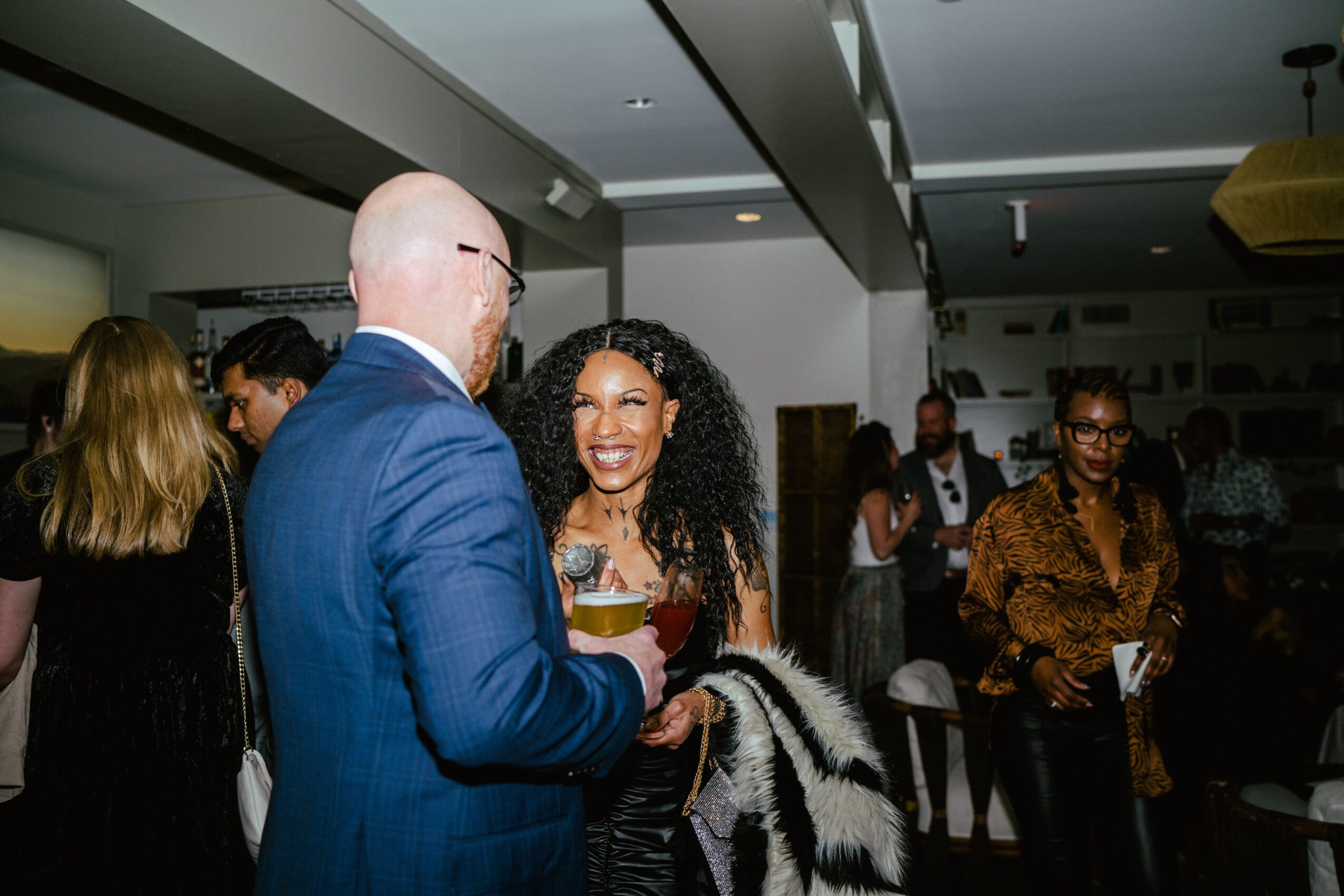 A woman with curly hair smiling and holding a drink, talking to a bald man in a blue suit at a social event.