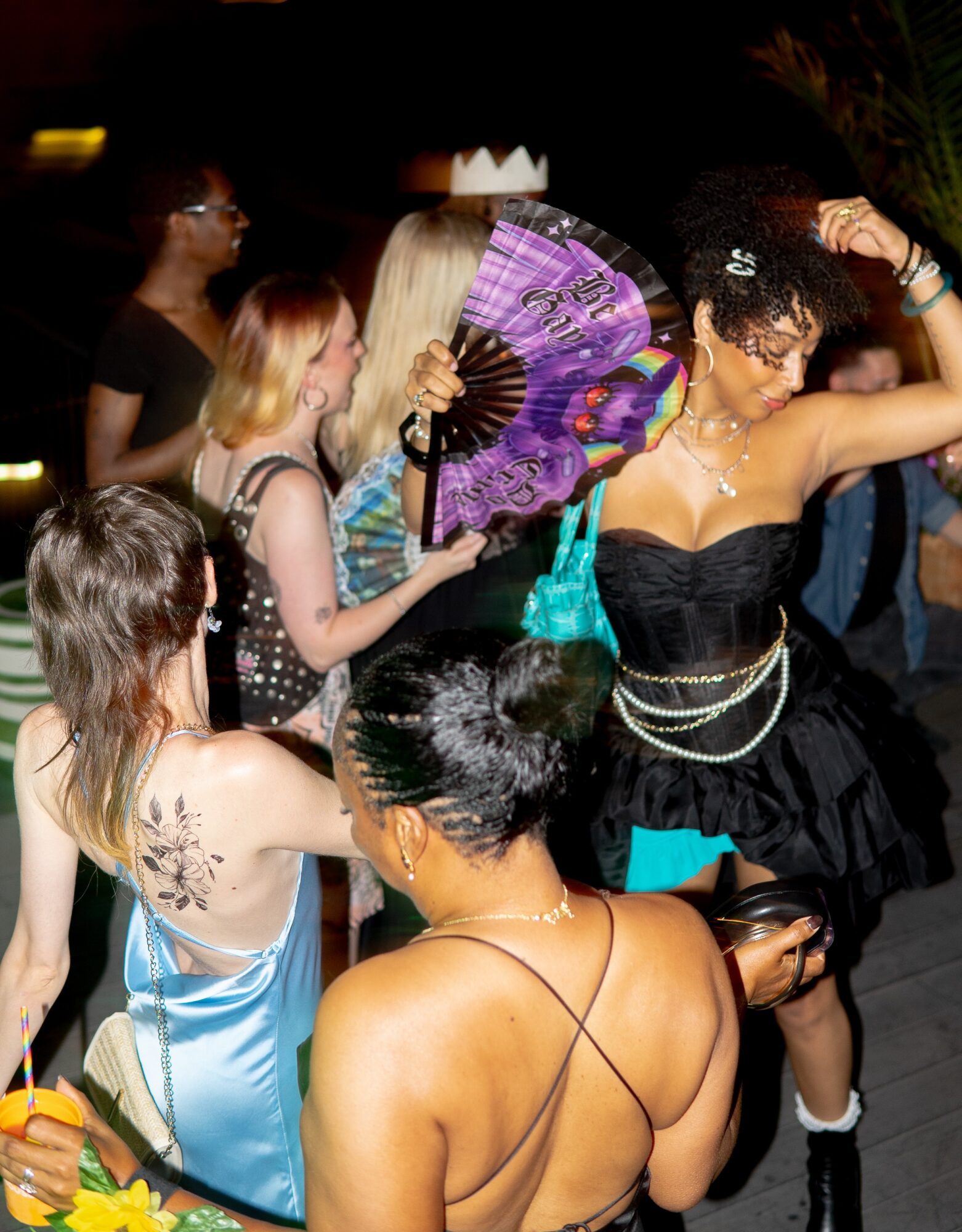 Group of women dancing at a party, one holding a purple fan, others in colorful dresses, in a dimly lit setting.