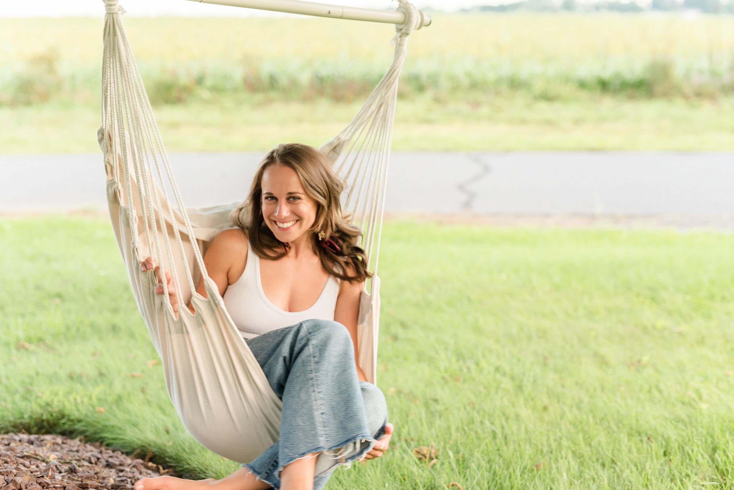 Woman sitting in a hammock outdoors, smiling, with green grass and a road in the background.