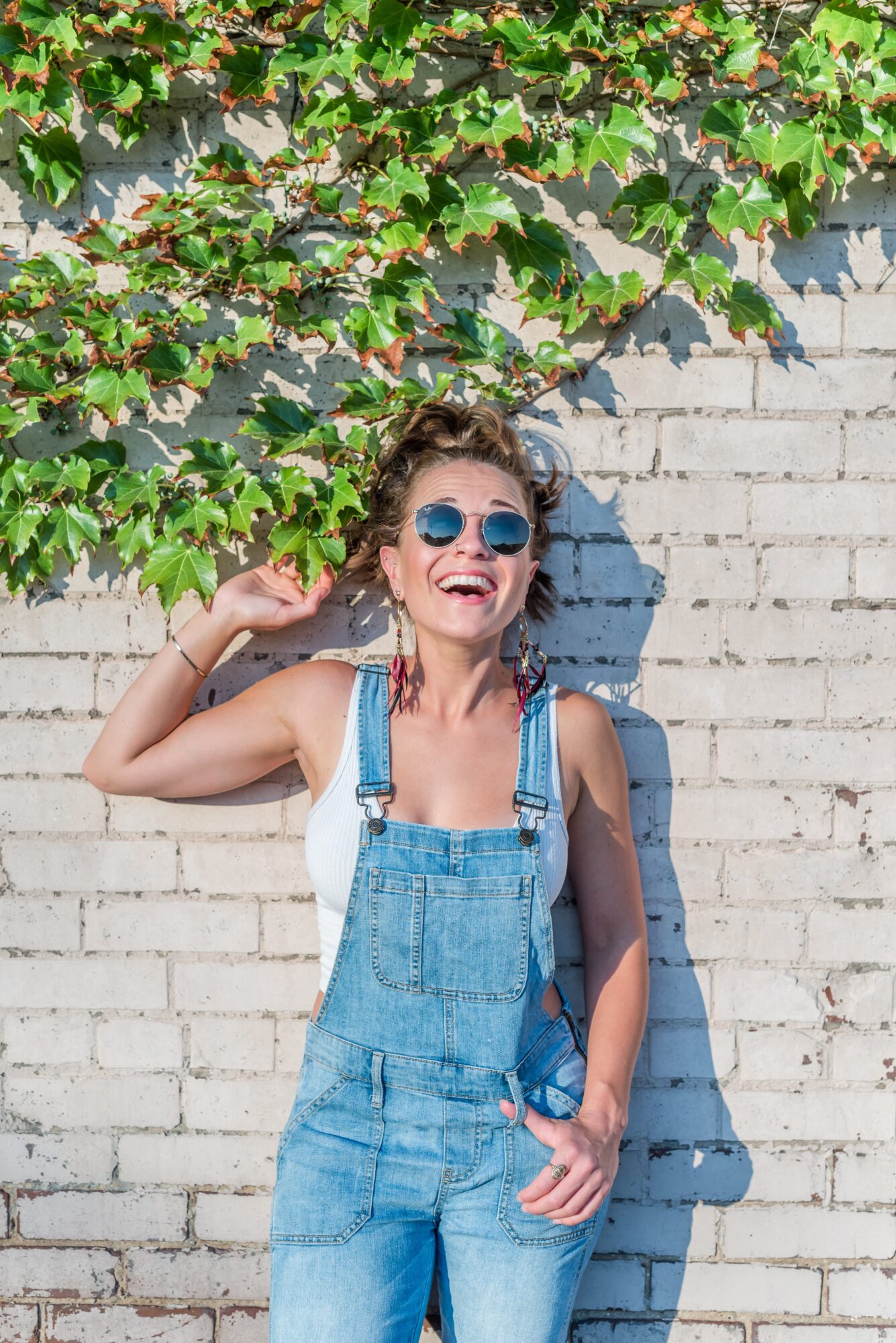 Young woman with sunglasses smiling, standing against a white brick wall with green leaves above her.