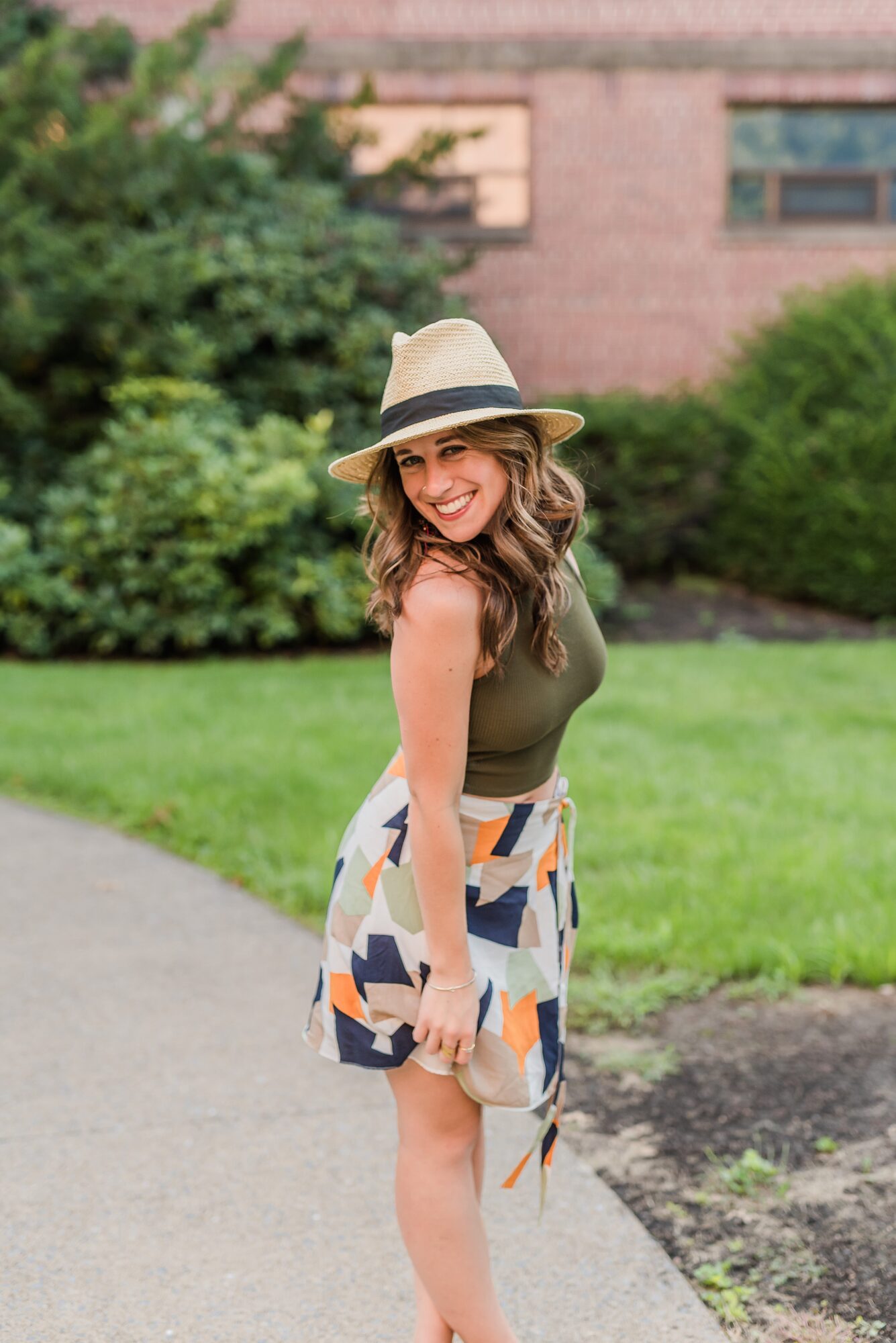 Smiling woman in a hat and patterned skirt standing outdoors on a grassy area with trees and a building in the background.
