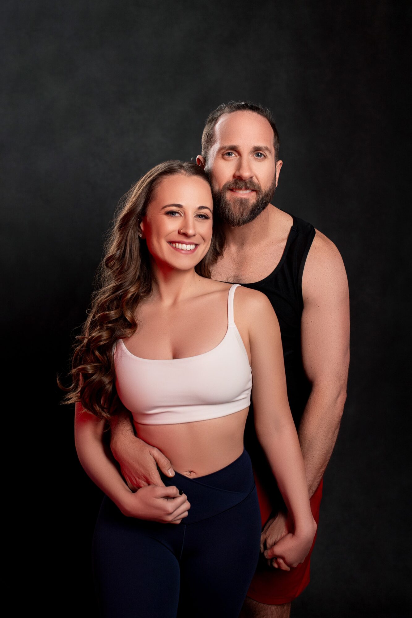 A smiling woman with long wavy hair and a man with a beard pose together against a dark background.