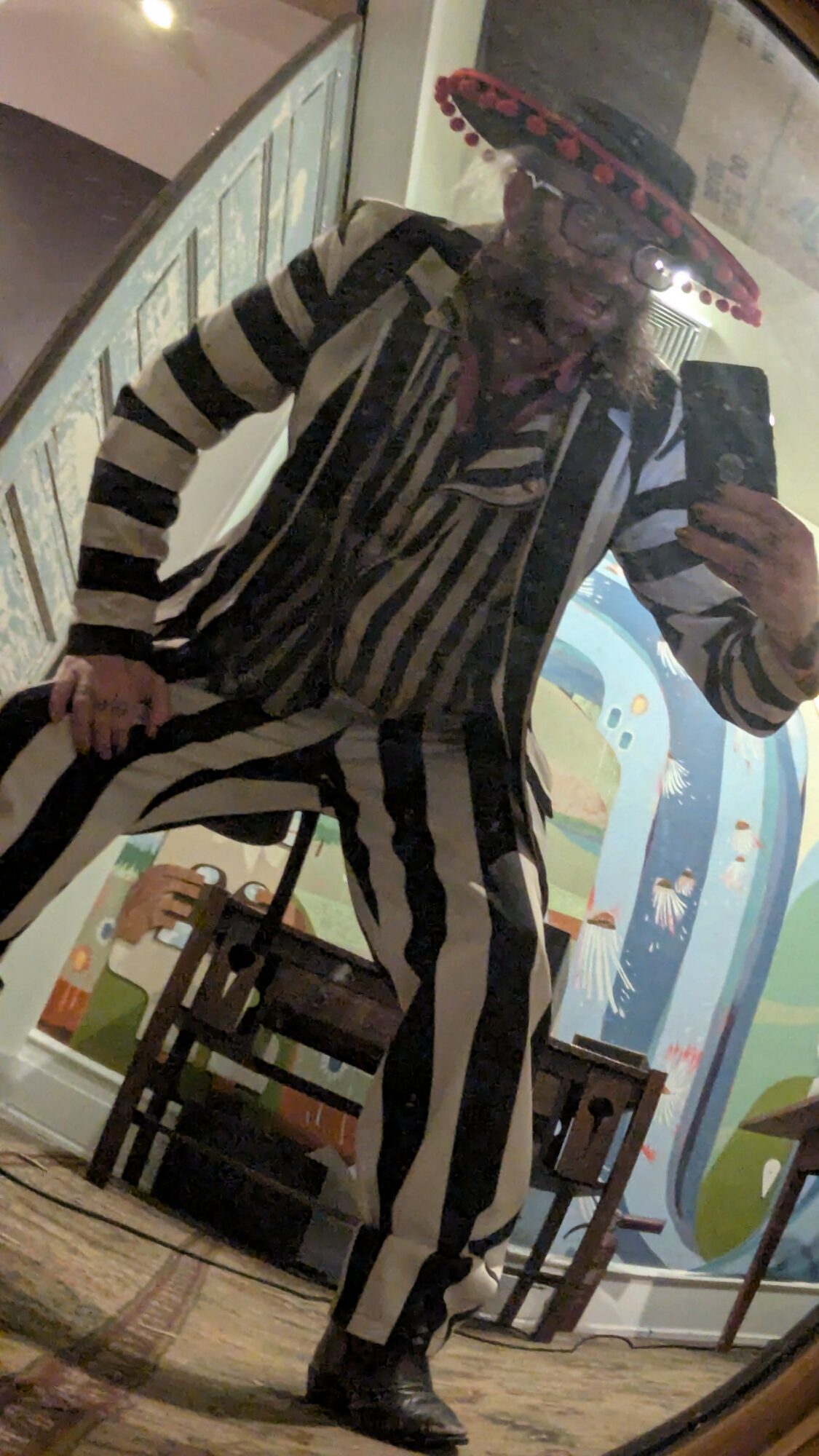 Person dressed as a clown in a striped costume, wearing a sombrero, taking a mirror selfie in a decorated room.