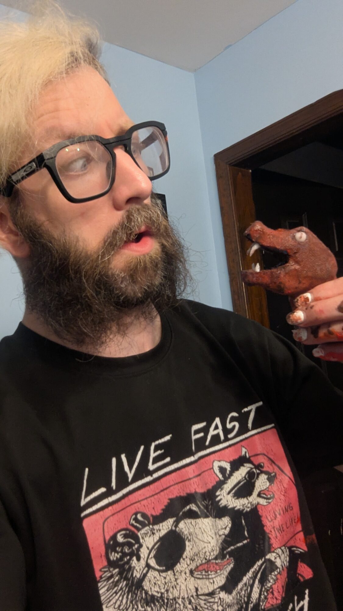 Person with glasses and a beard looking to the side, holding a small object in their hand, indoors.