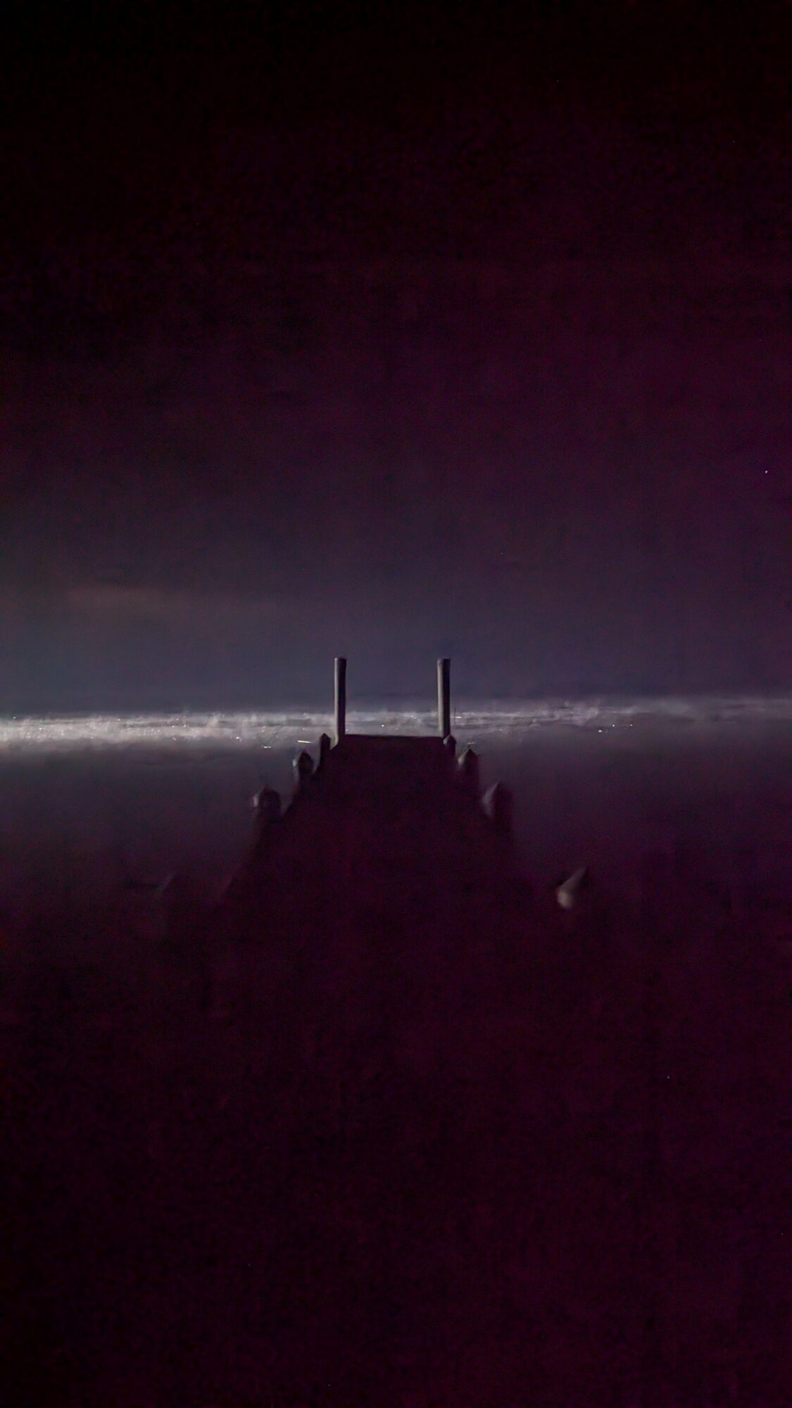 Dark scene with a silhouette of a structure or pier extending into water, horizon line visible, sky dark with faint light.