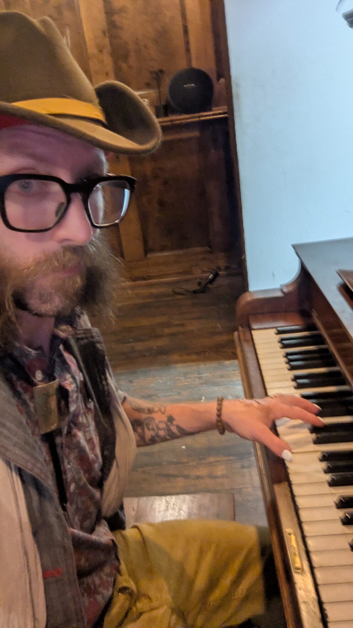 Person wearing glasses and a hat playing piano in a room with wooden walls and floor.