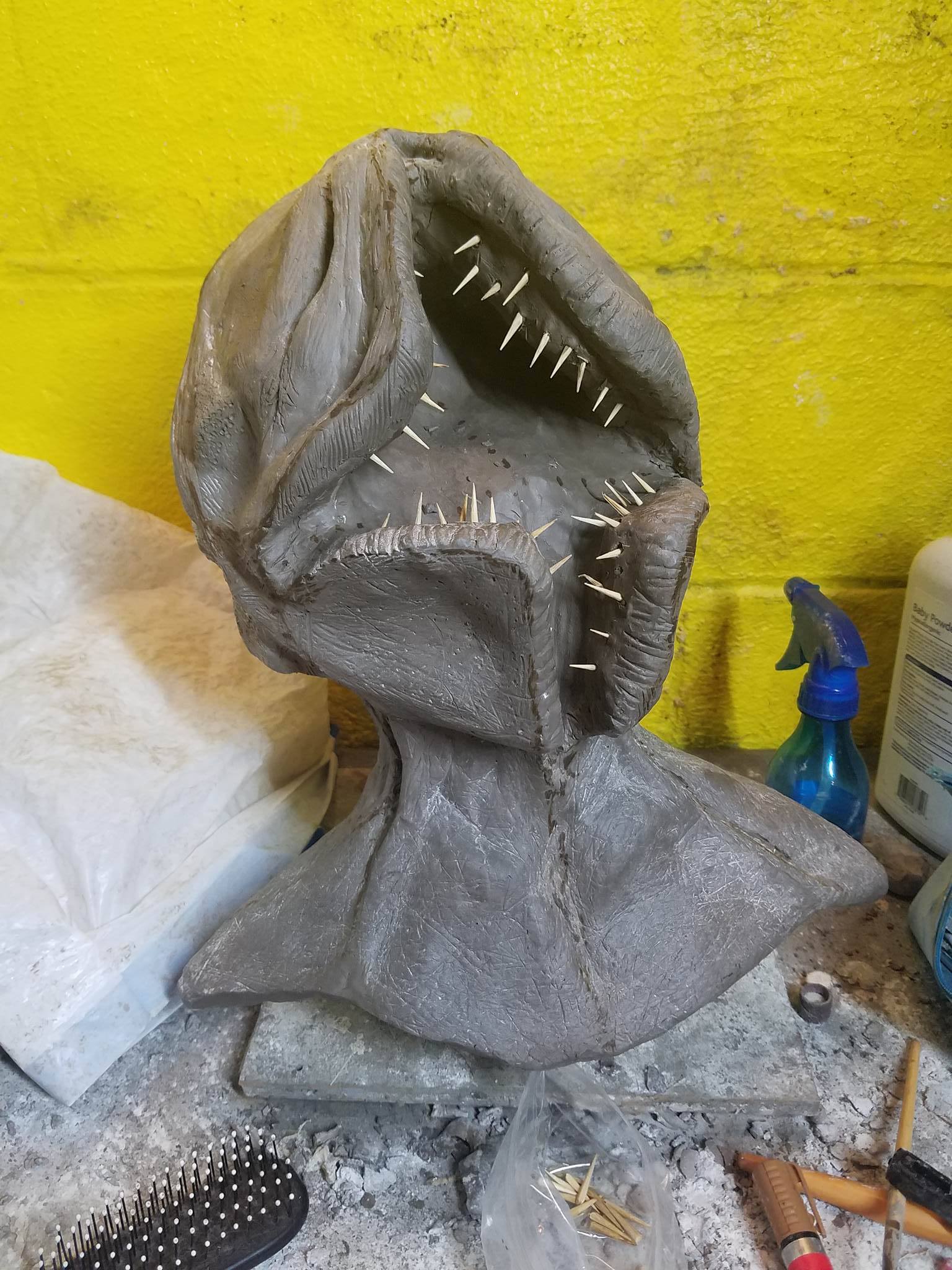 Sculpture of a creature with an open mouth, large head, and textured surface, set against a yellow wall.