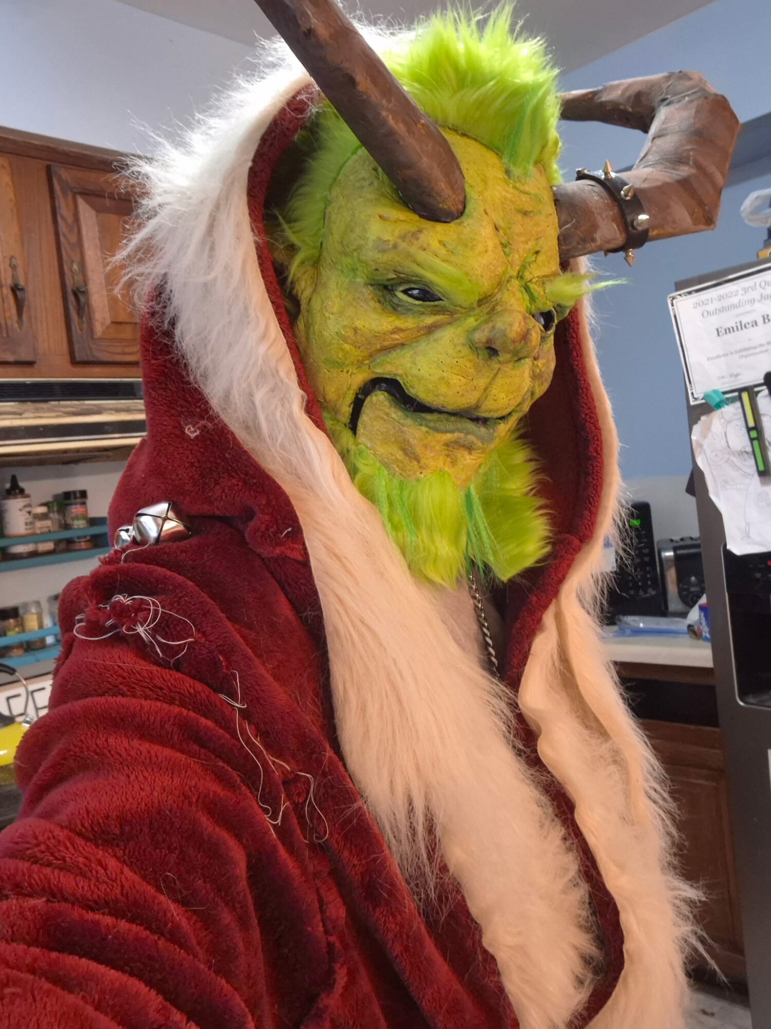 Person in a green demon mask with horns, yellow face, and long white hair, wearing a red hooded cloak, indoors.