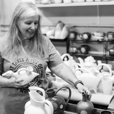 Woman smiling while pouring tea into a teapot in a kitchen, surrounded by teapots and cups, black and white photo.