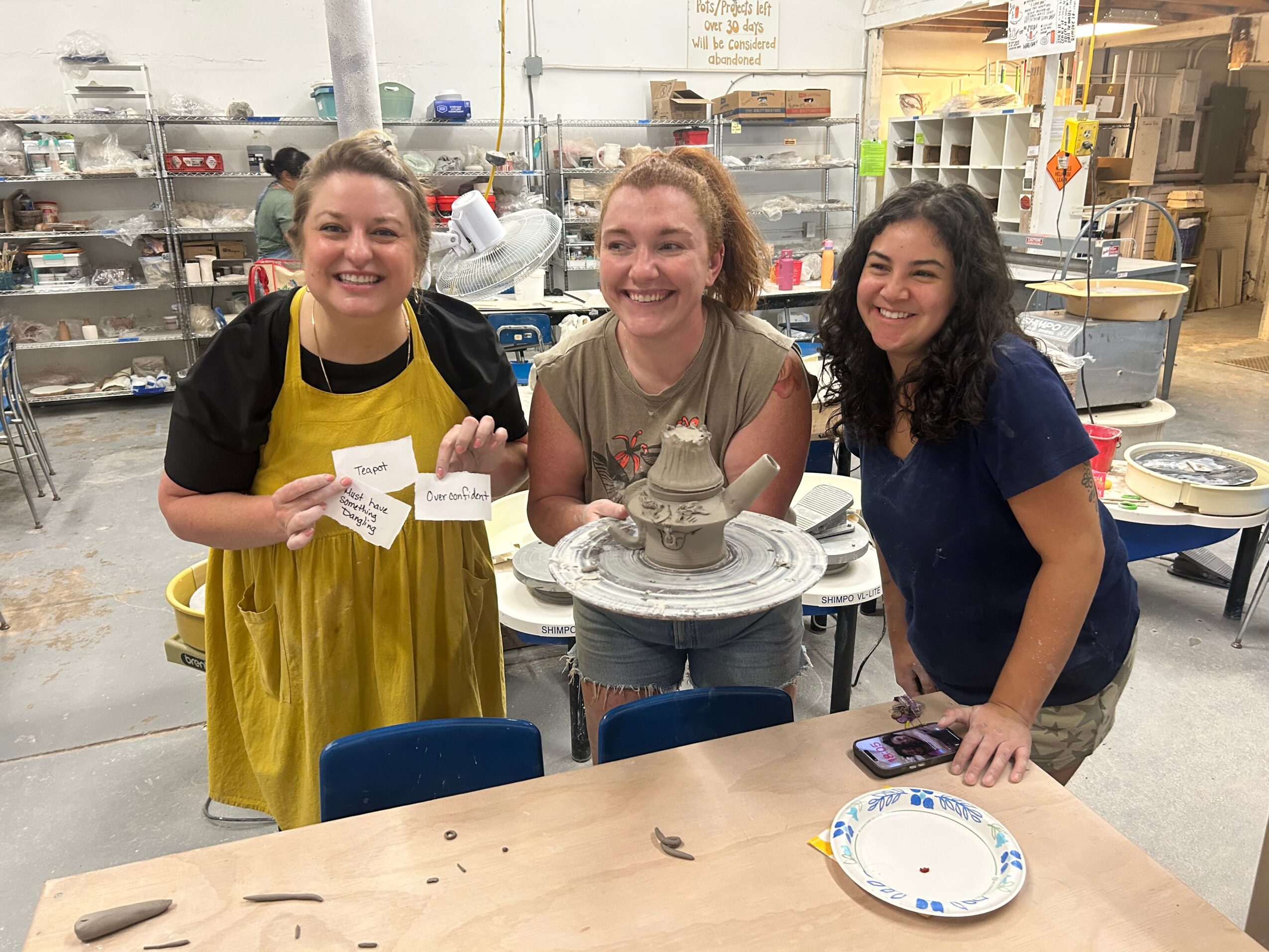 Three women smiling and posing in a workshop with pottery equipment, standing behind a table with a plate and phone.