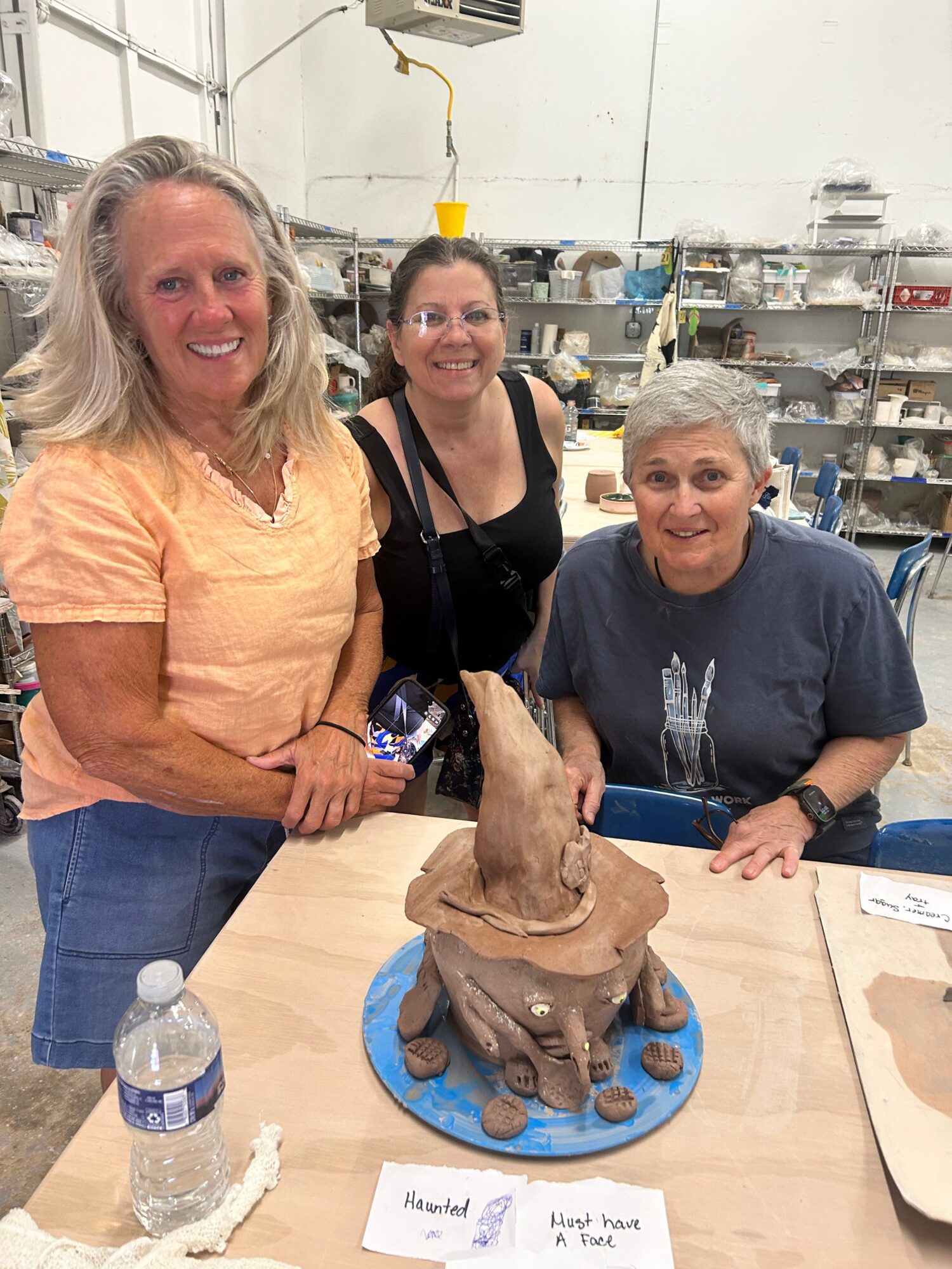 Three people around a table with a clay sculpture, water bottle, and papers in a workshop setting.