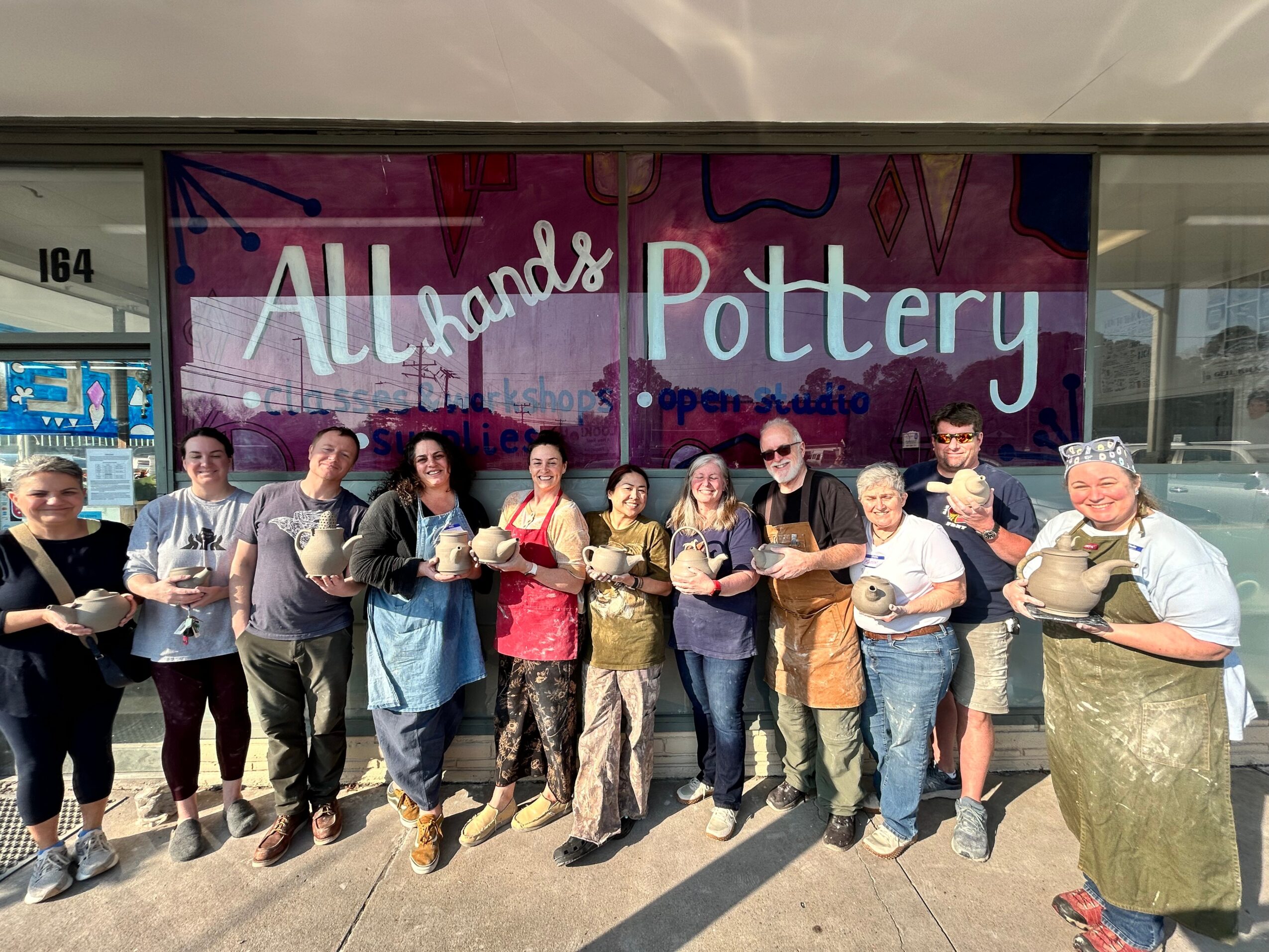 Group of people holding pottery items outside a store named All Hands Pottery, with a purple sign and large window, sunny day.